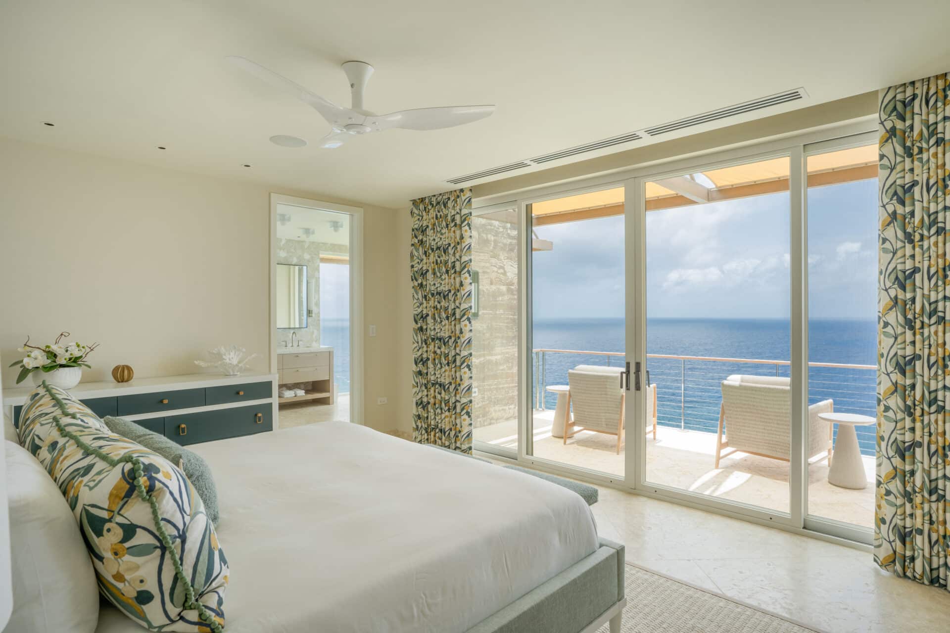 A serene bedroom in Villa Quintessa features a large bed with floral-patterned pillows and matching curtains. The room opens to a balcony through sliding glass doors, offering stunning ocean views. A stylish dresser and a ceiling fan complete the minimalist decor.