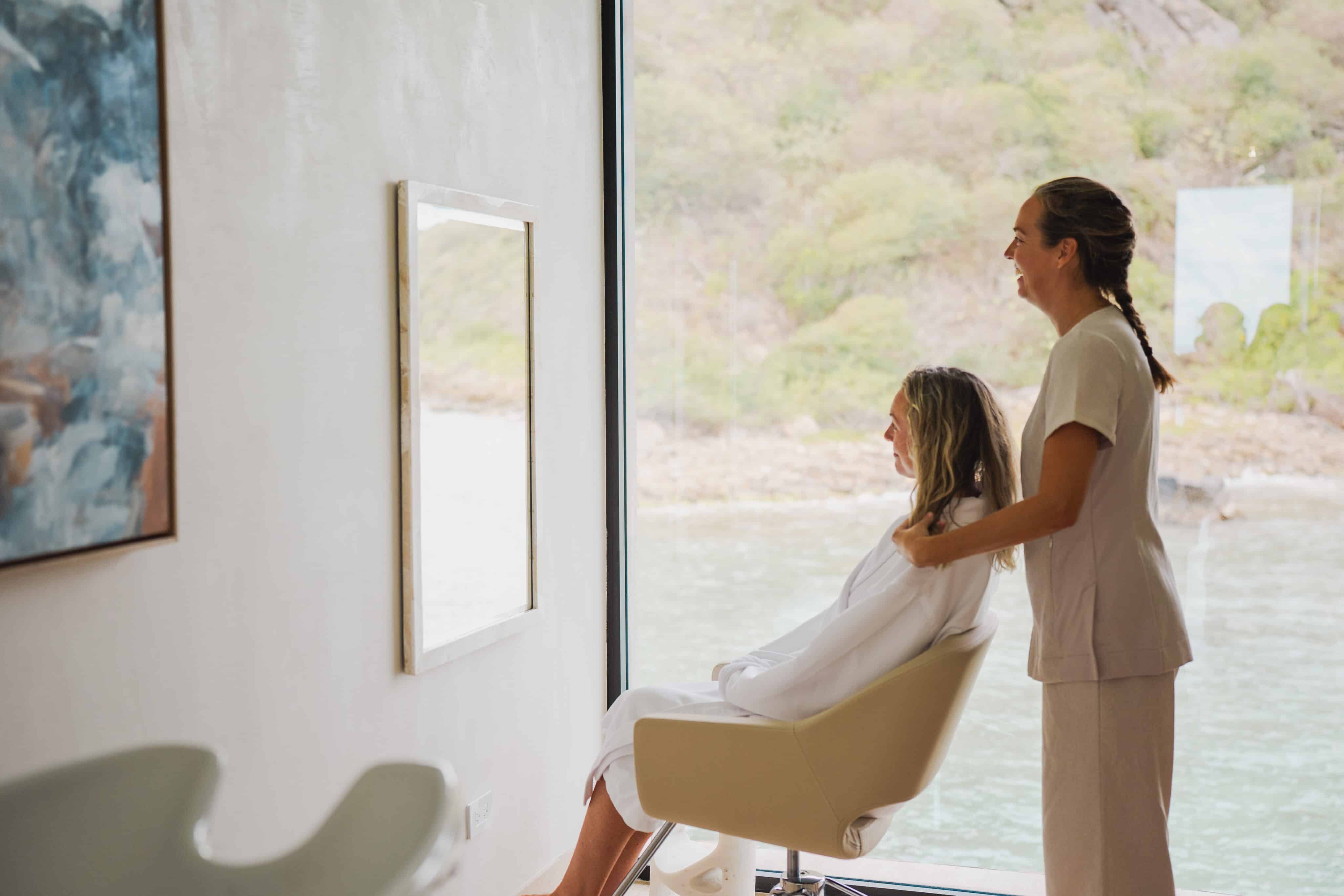 A woman sits in a salon chair at the Oil Nut Bay spa, facing a scenic water view, while another woman stands behind her, gently massaging her shoulders. The bright, modern room features art on the wall.
