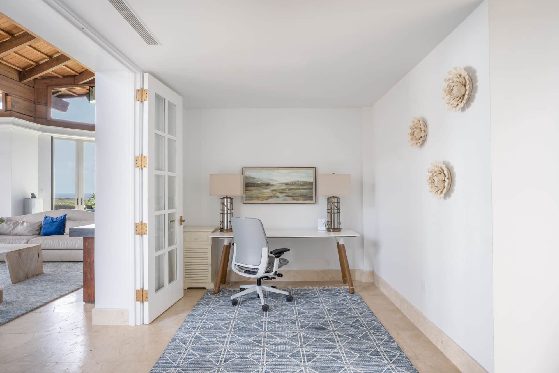 A minimalist home office in a villa, featuring a sleek white desk with a modern office chair, flanked by two beige lamps. A landscape painting hangs above the desk. The room has white walls, a geometric-patterned rug, and three decorative wall pieces. Wings of the adjoining living room are visible.