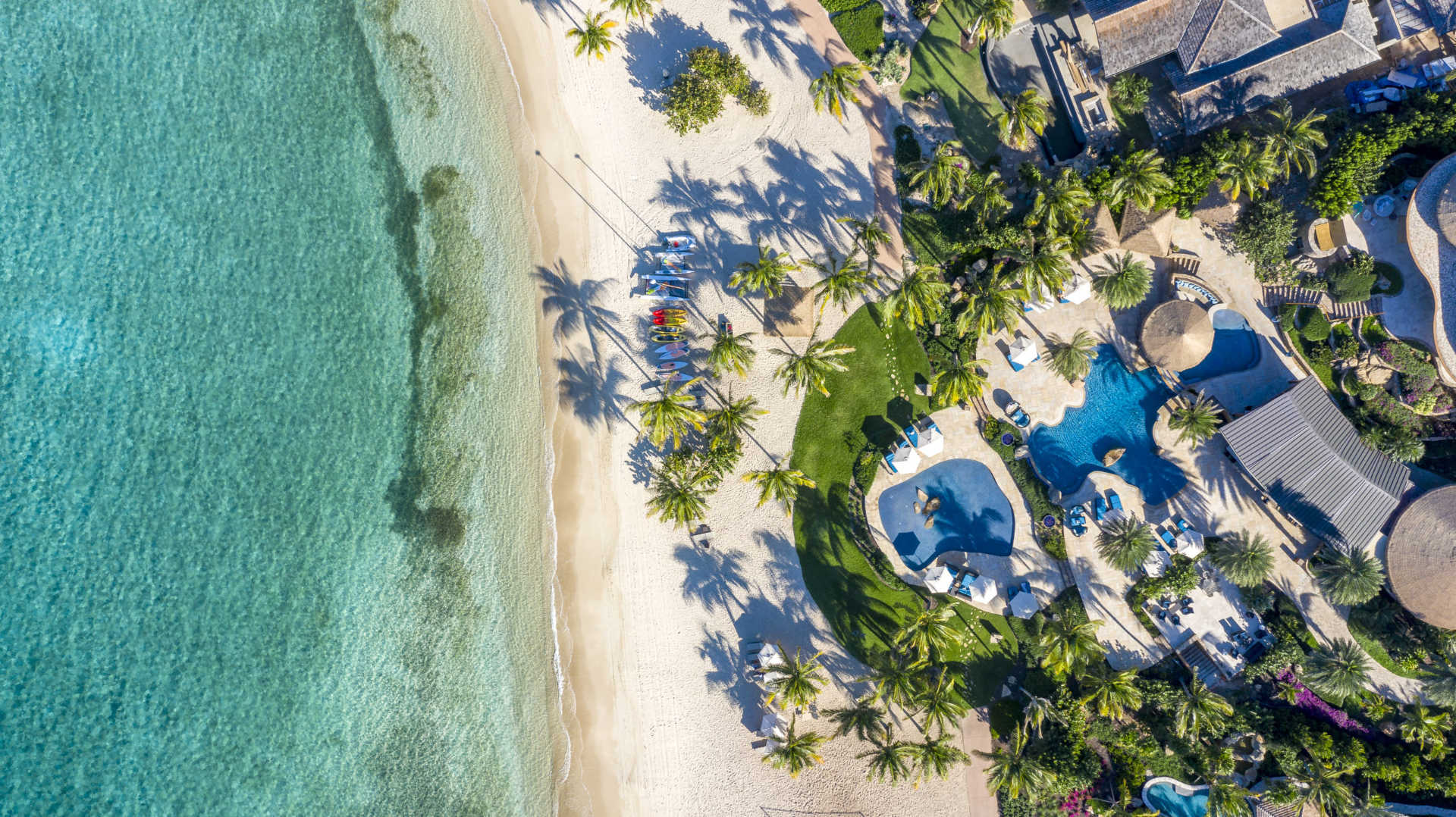 Aerial view of a tropical beach with clear turquoise water, white sand, and several people gathered near sailboats. The shoreline is adorned with palm trees and lush greenery. Buildings and swimming pools are visible in a resort area next to the beach—an idyllic spot for Caribbean vacations.
