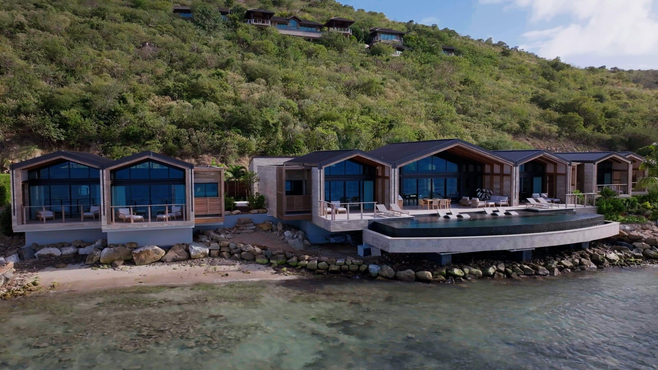 Modern hillside villa with large glass windows and an infinity pool overlooking a serene beachfront, reminiscent of a James Bond getaway. The house is set against lush greenery, with a clear blue sky above, creating an atmosphere worthy of a Royale experience.