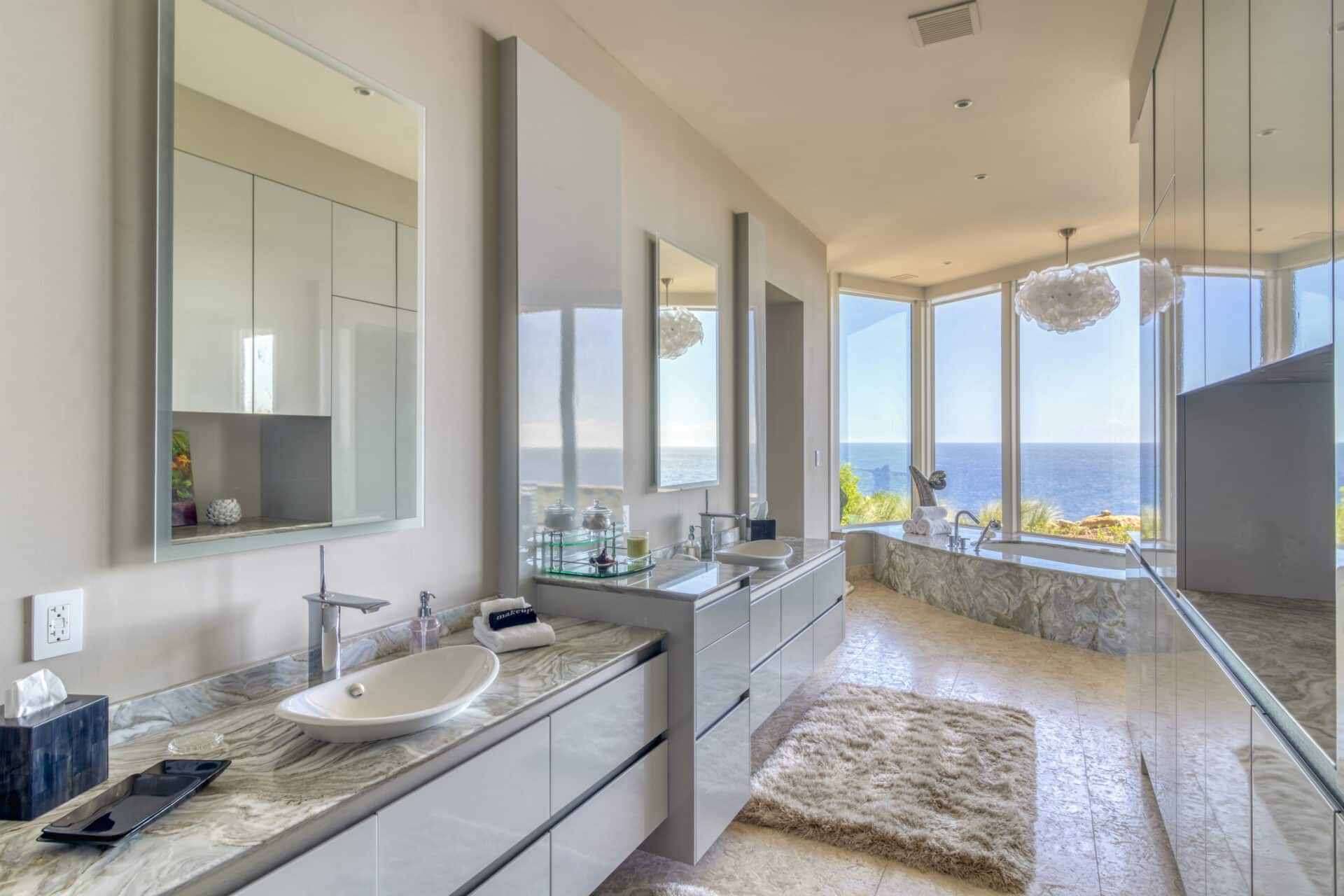 A modern bathroom in Halo Villa features a marble countertop, dual sinks, wall-mounted mirrors, and minimalistic decor. A large window offers an ocean view. The room is bright with natural light, and next to the window sits a bathtub with a fluffy rug on the marble floor.