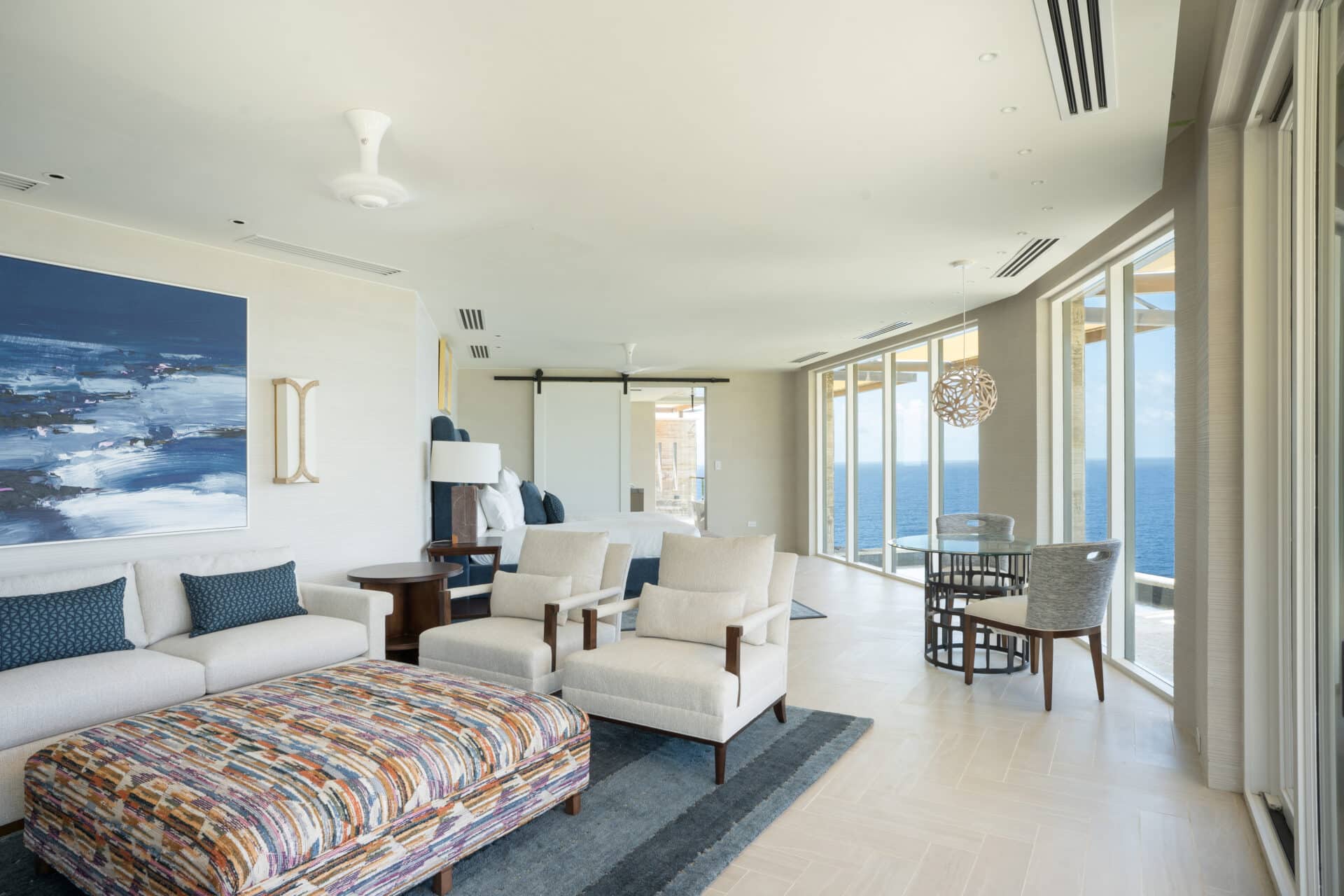 A modern living room in Villa Quintessa with white furniture and blue accents, featuring a sofa, two armchairs, and a colorful ottoman. Large windows offer an expansive ocean view. The room includes a round dining table with chairs and a sliding barn door leading to another area.