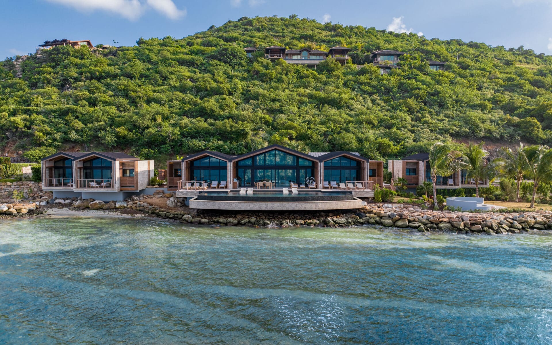 A modern, luxury ocean villa with large glass windows is set against a lush, green hillside. Surrounded by palm trees and overlooking clear, calm waters, OV1 creates a serene and tropical atmosphere.