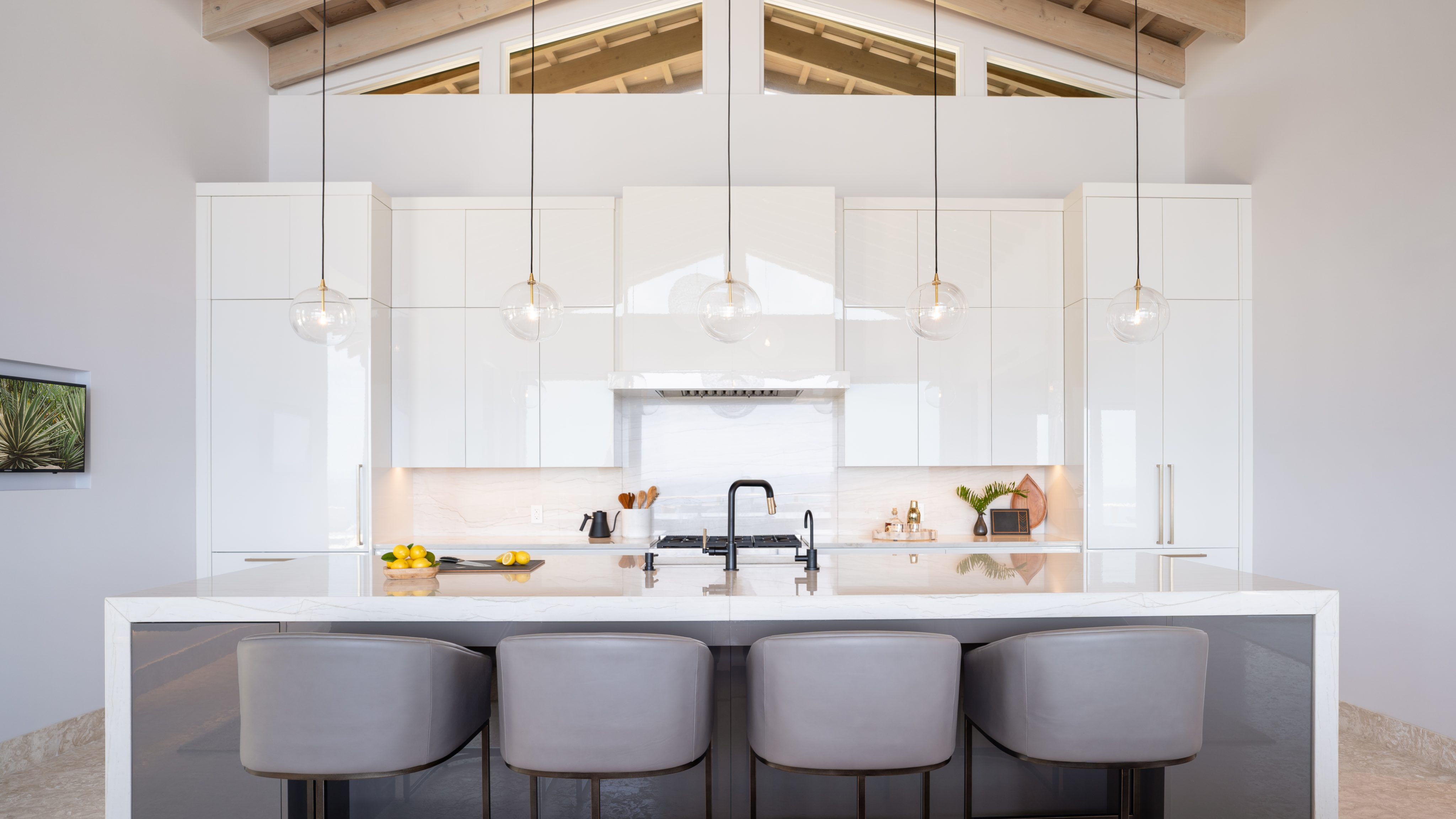 A modern villa kitchen features a sleek white design with high ceilings and open beams. A large island with a marble countertop and four gray barstools occupies the center. Pendant lights hang above the island, resembling graceful hummingbirds in flight. The backdrop includes cabinets, a stove, and a range hood.