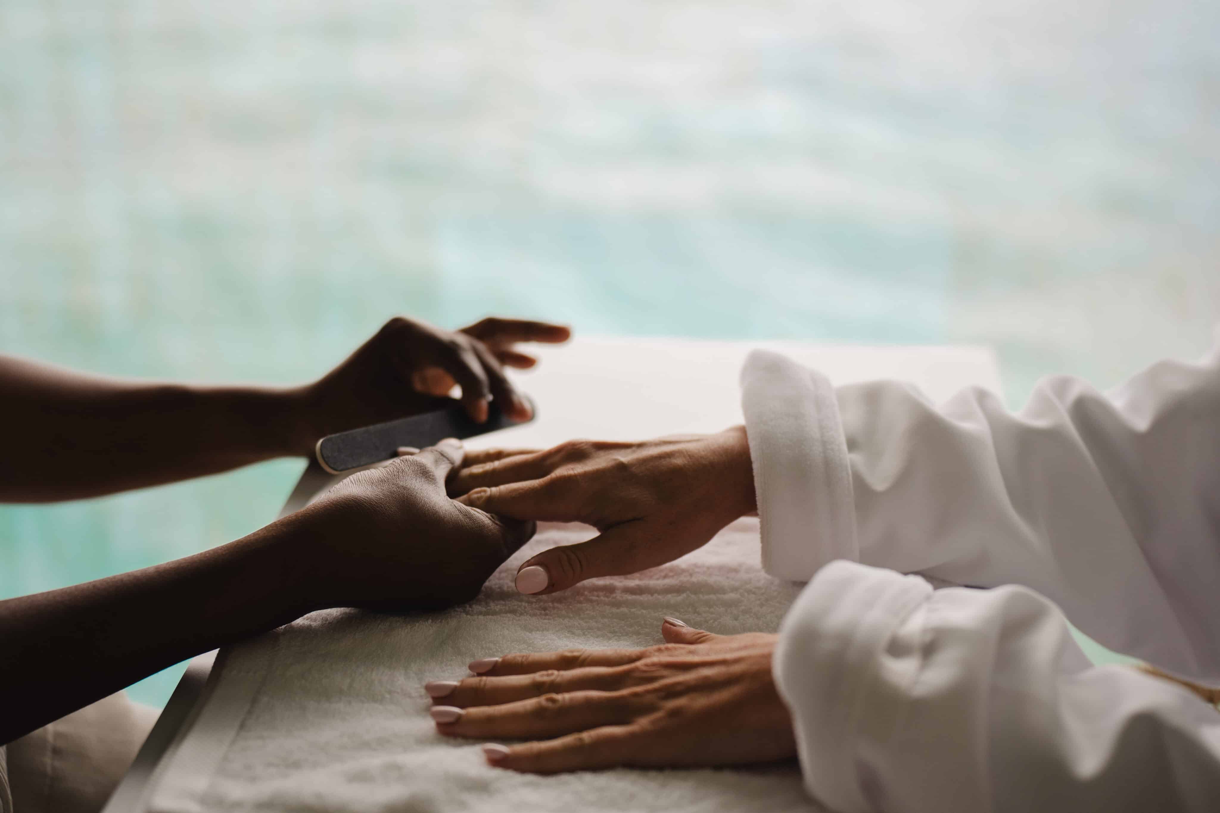 A person enjoys a manicure at the Oil Nut Bay spa, as another files their nails over a white towel. Both are dressed in white robes, with a softly blurred background enhancing the serene atmosphere.