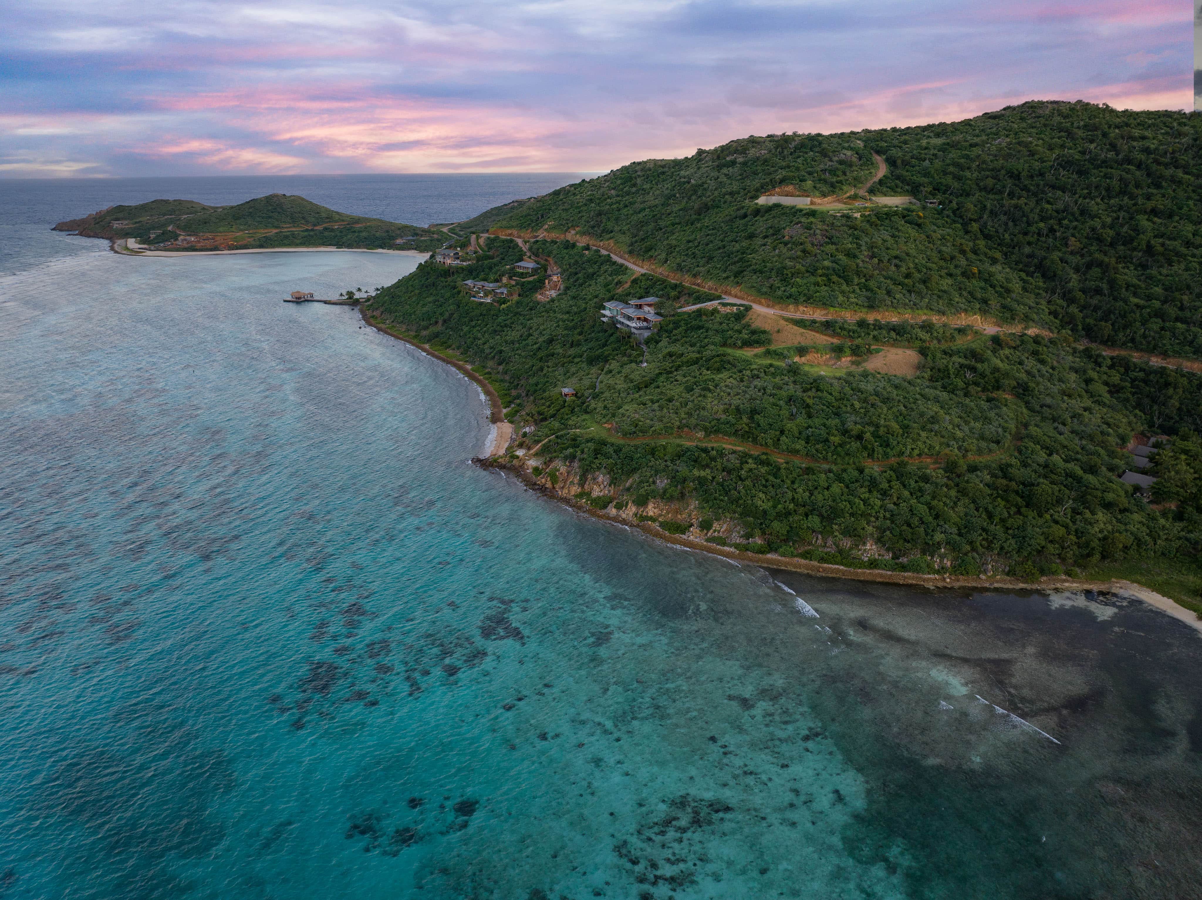 Aerial view of a coastal landscape with lush green hills and clear turquoise waters. The E15 road elegantly winds along the coastline. The sky, a mix of pink and blue hues, suggests either sunrise or sunset. Estate homesites dot the area, and small islands are visible in the distance.