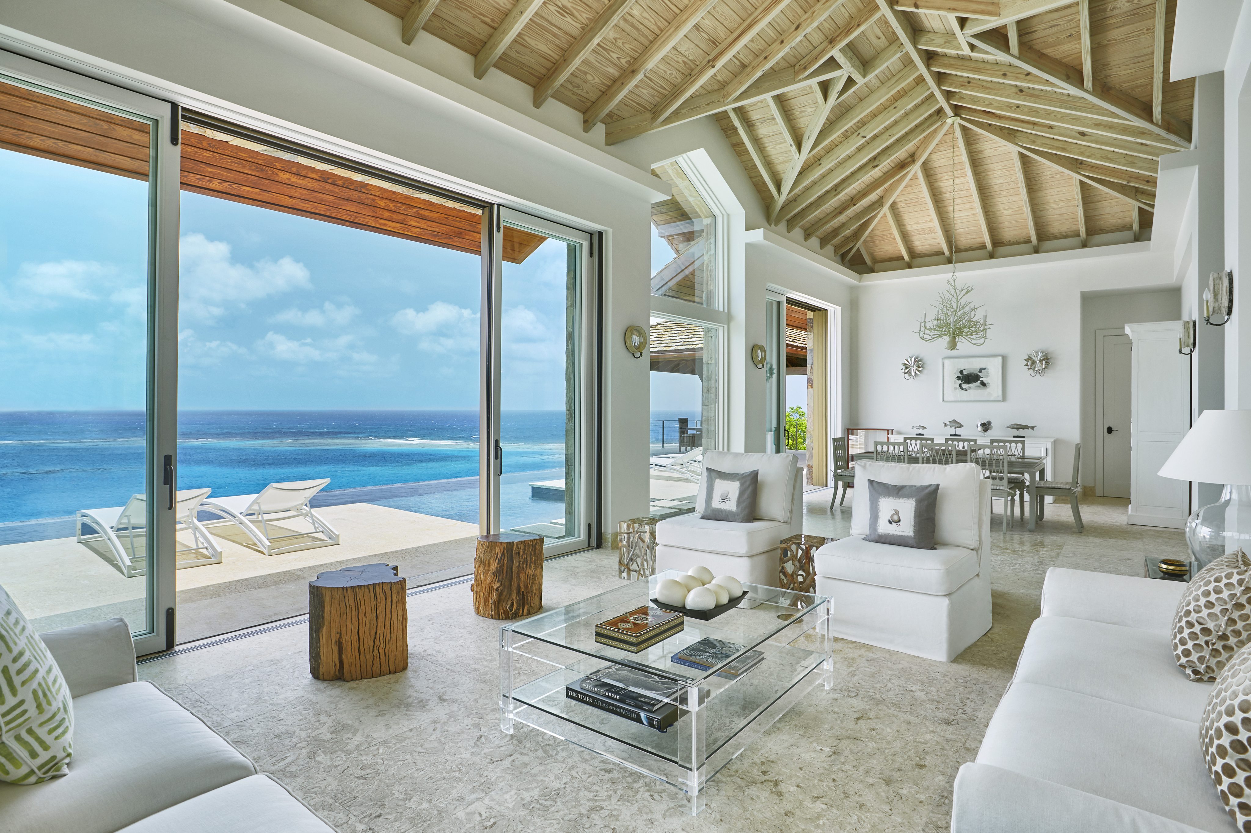 A modern, open-concept living room at Reef House features large sliding glass doors offering a stunning ocean view. The room includes white furniture, a glass coffee table, and wooden accents. Outdoor patio chairs are visible by the pool, and the ceiling has exposed wooden beams.