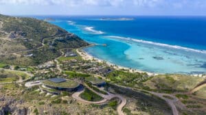 Aerial view of a coastal area in the British Virgin Islands featuring a luxury resort for sale close to a turquoise, sandy beach. The landscape includes lush green hills and winding roads, leading to well-designed buildings with modern architecture by Lion Heart Home. The ocean stretches out to small islands in the distance.