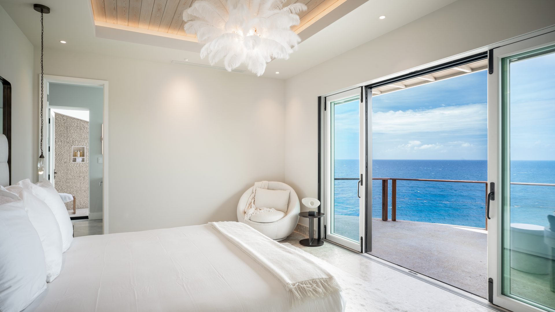 A modern villa bedroom features a large sliding glass door opening to a balcony with a stunning ocean view. The room has a white bed, a cozy round chair, and a unique feathered light fixture reminiscent of a hummingbird. The décor is minimalist, with light-colored walls and ceiling.
