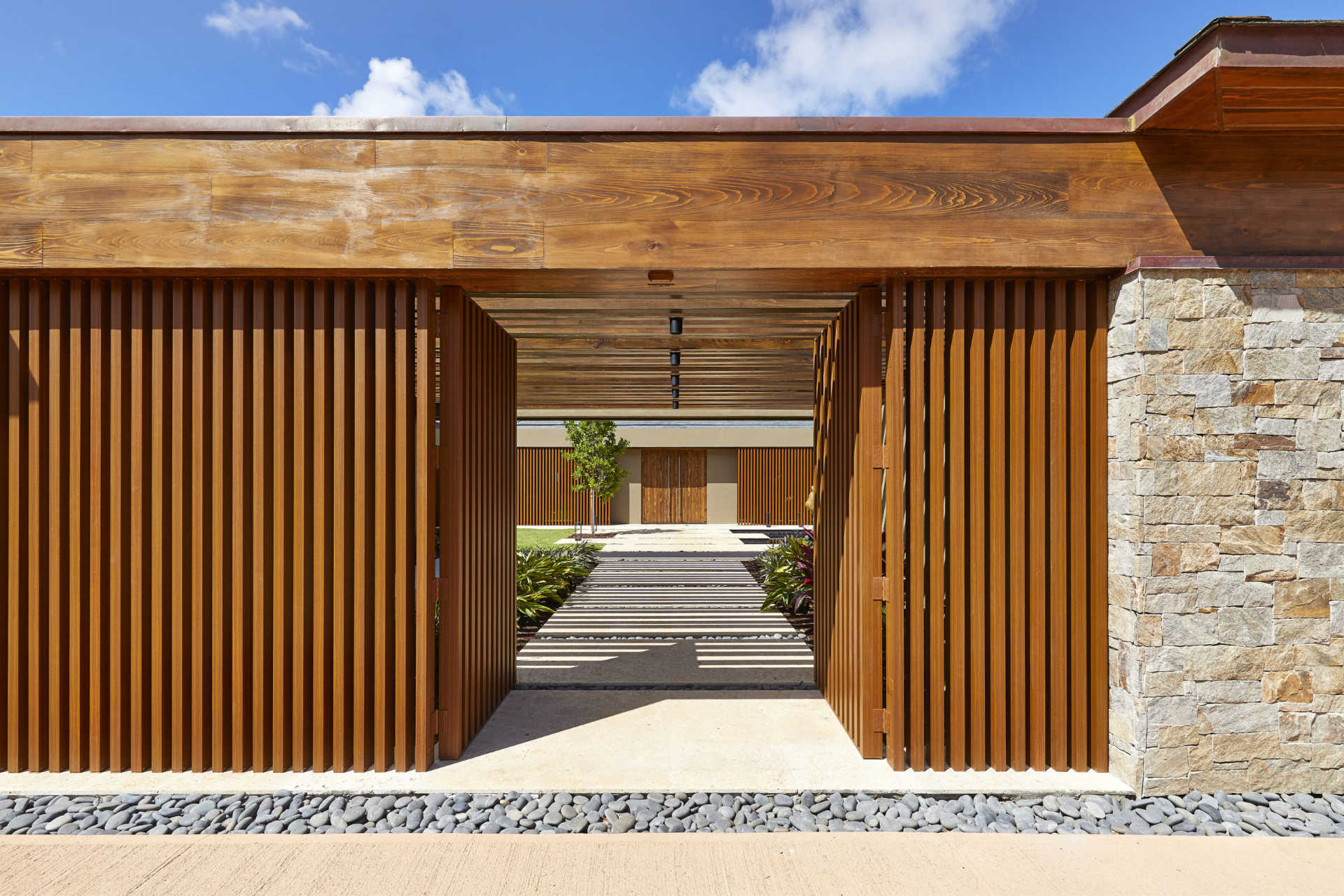 Modern architectural entrance featuring vertical wooden slats with a stone wall on the right. The structure leads to a pathway lined with trees and plants, extending towards a villa in the background under a partly cloudy sky.