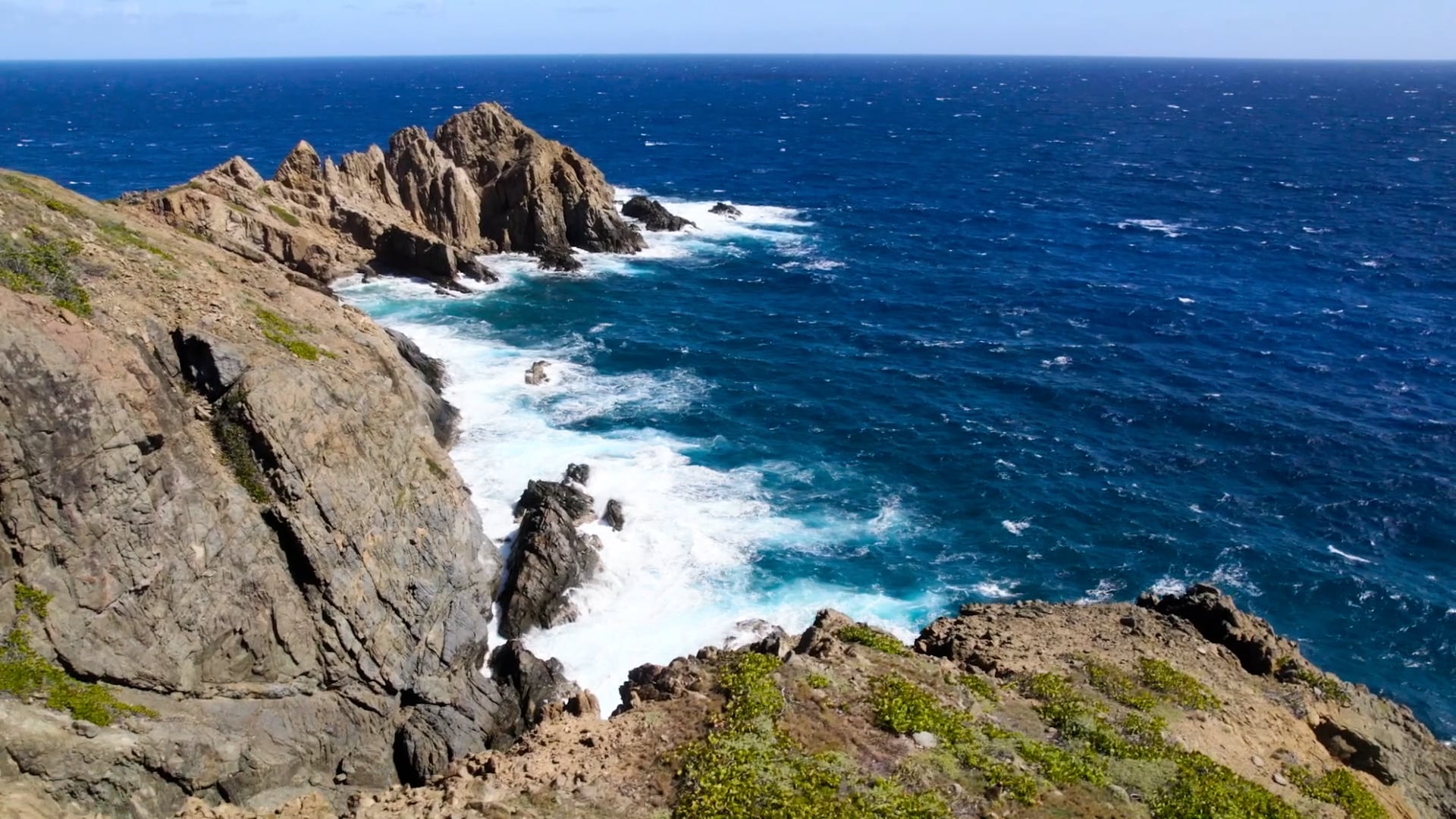 A rocky coastline with jagged cliffs and crashing ocean waves under a clear blue sky, reminiscent of The Point. The deep blue sea churns white foam against the rocks, while sparse greenery clings to the cliff edges near this picturesque homesite.
