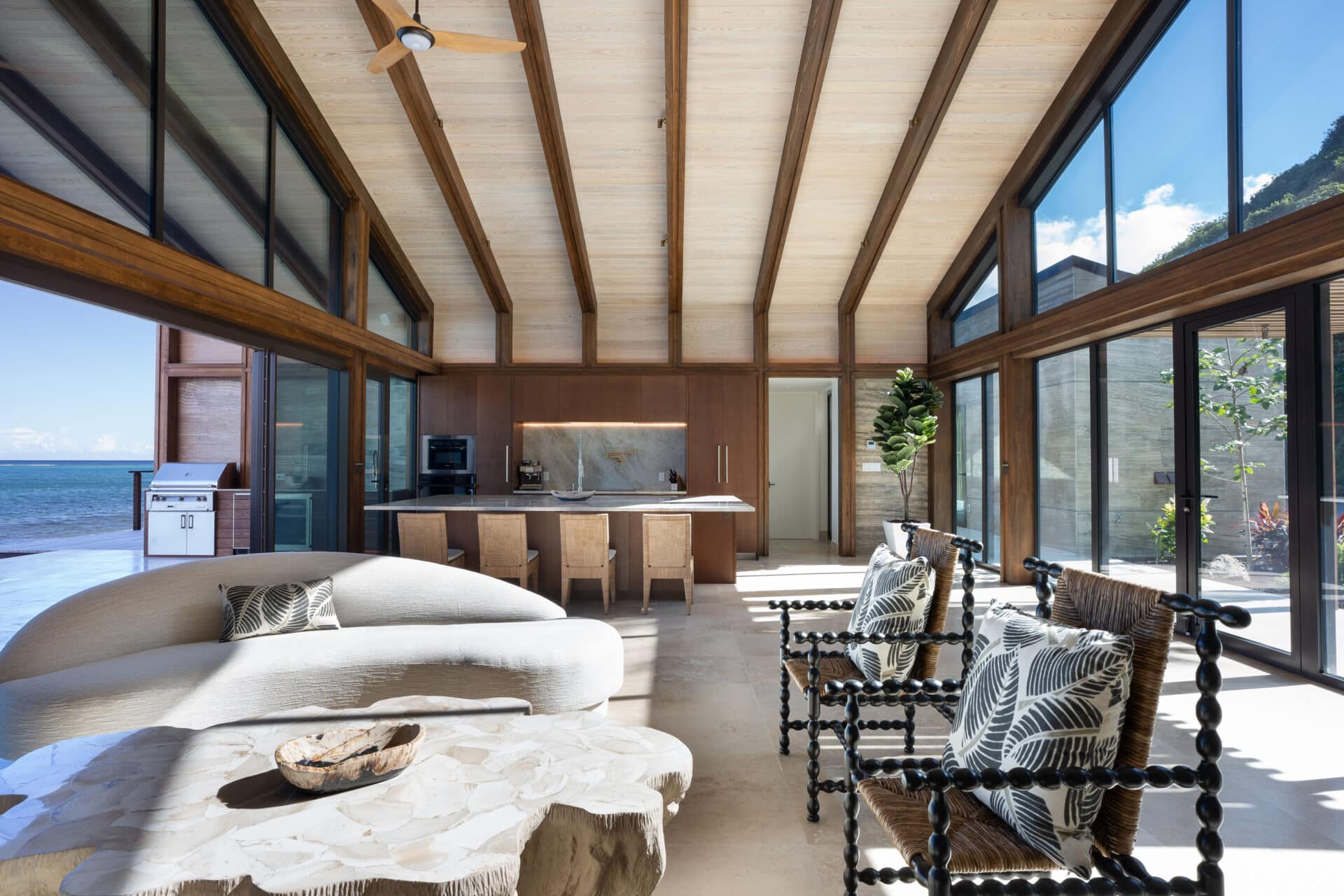 A spacious, modern living room in Ocean Villa boasts large windows that overlook the ocean. The room features a curved sofa, two armchairs, and a stone table. High wooden ceilings and a minimalist kitchen enhance the airy ambiance.