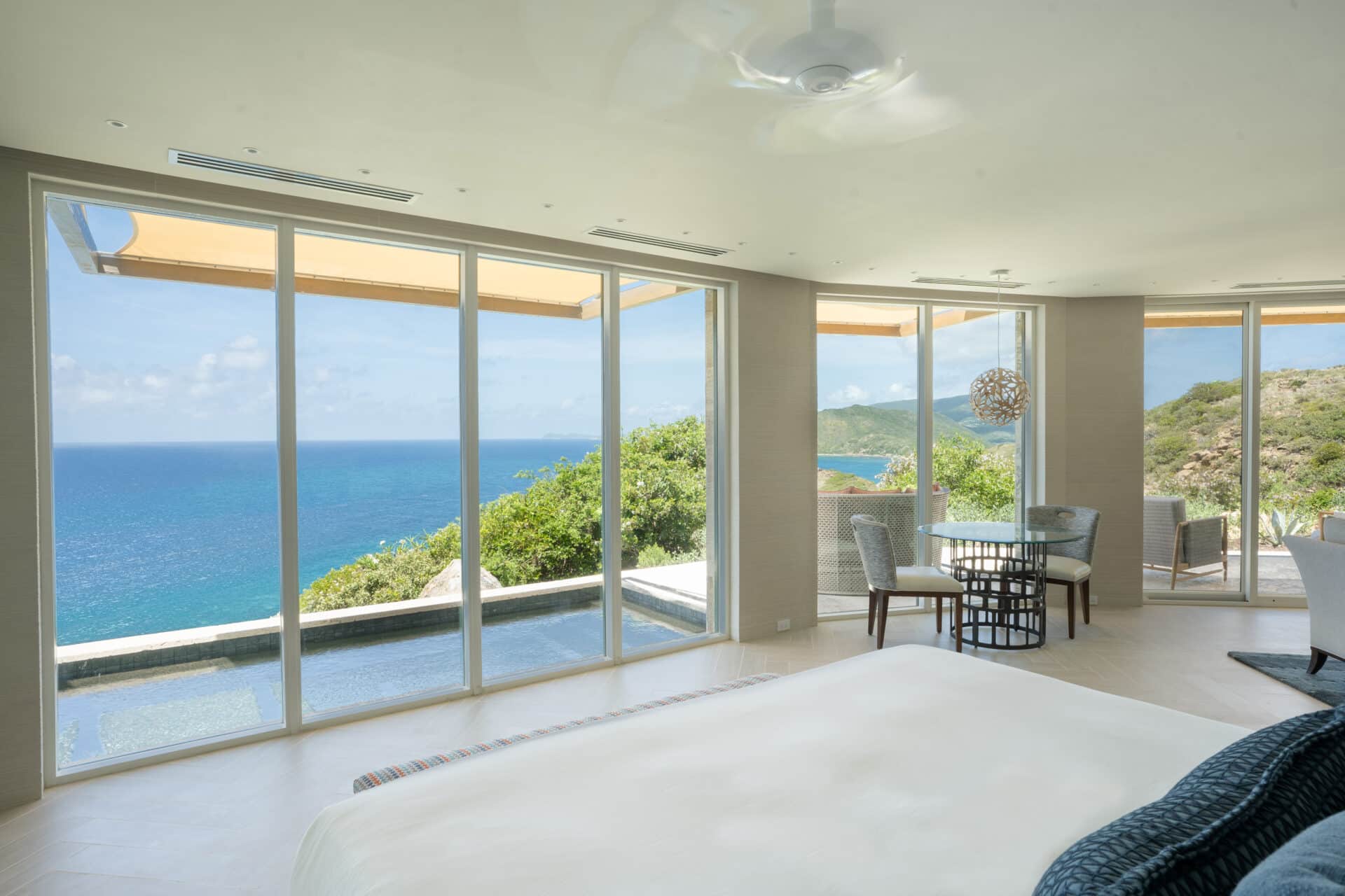 Spacious bedroom with large windows offering a panoramic view of the ocean and surrounding greenery at Quintessa Villa. The room features a white, neatly made bed and a small dining area with a round table and four chairs. Blinds hang above the windows, and a ceiling fan is visible in this beautiful villa rental.