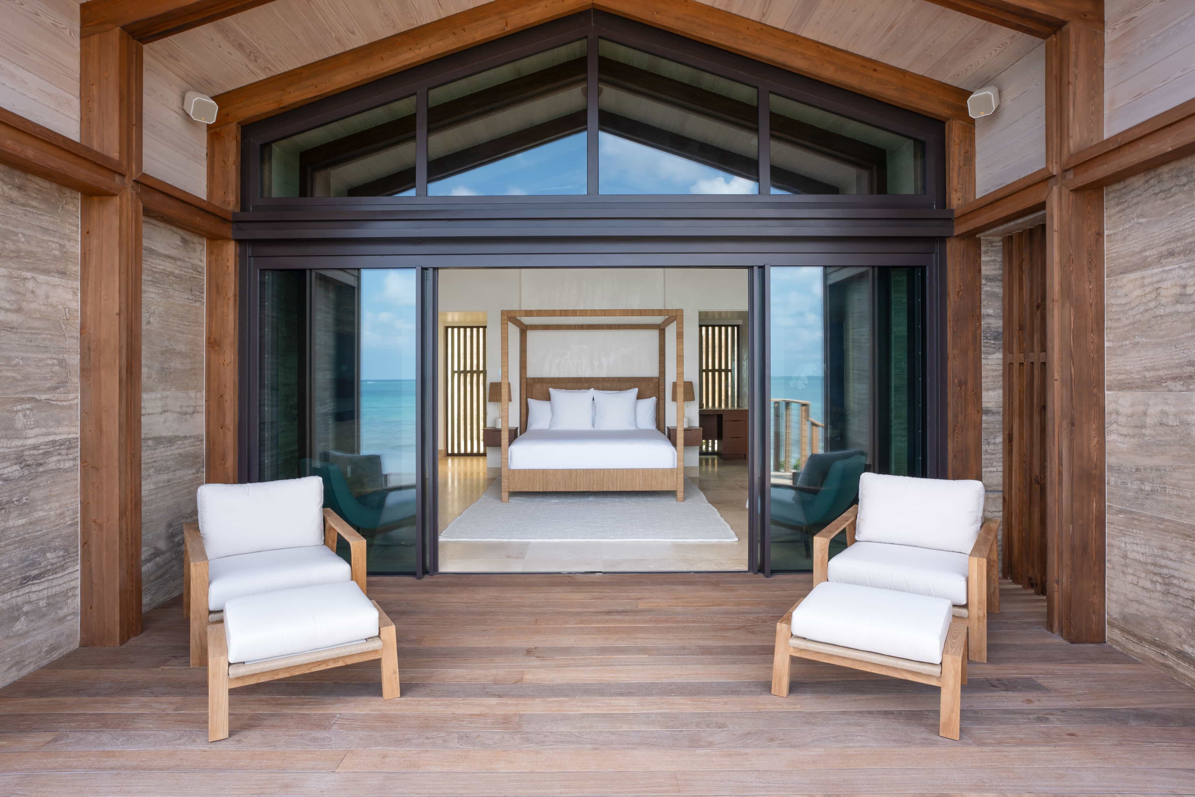 Luxurious Ocean Villa with a modern design, featuring a wooden deck. Two white cushioned chairs face a spacious bedroom with a four-poster bed, large windows providing ample natural light, and an exquisite view of the ocean.
