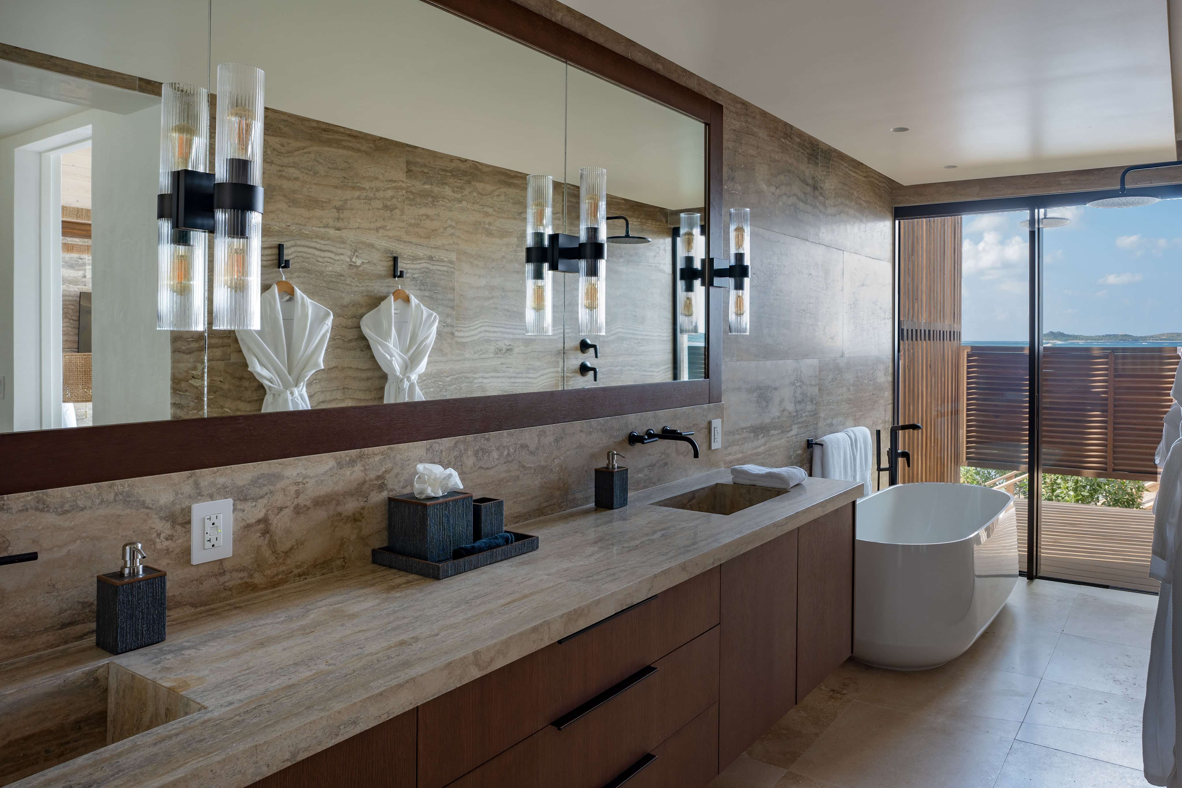 Luxurious bathroom at Ocean Villa with dual sinks, a large mirror, and elegant lighting. Features a standalone bathtub near a window showcasing a scenic view. The decor includes marble surfaces and wooden accents, creating a modern and serene atmosphere in OV1 style.