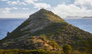 A narrow, green hill rises sharply, with a few people standing at its peak. The expansive ocean and a partly cloudy sky create a serene backdrop in this luxurious escape at Oil Nut Bay. Distant islands of the British Virgin Islands are visible on the horizon.