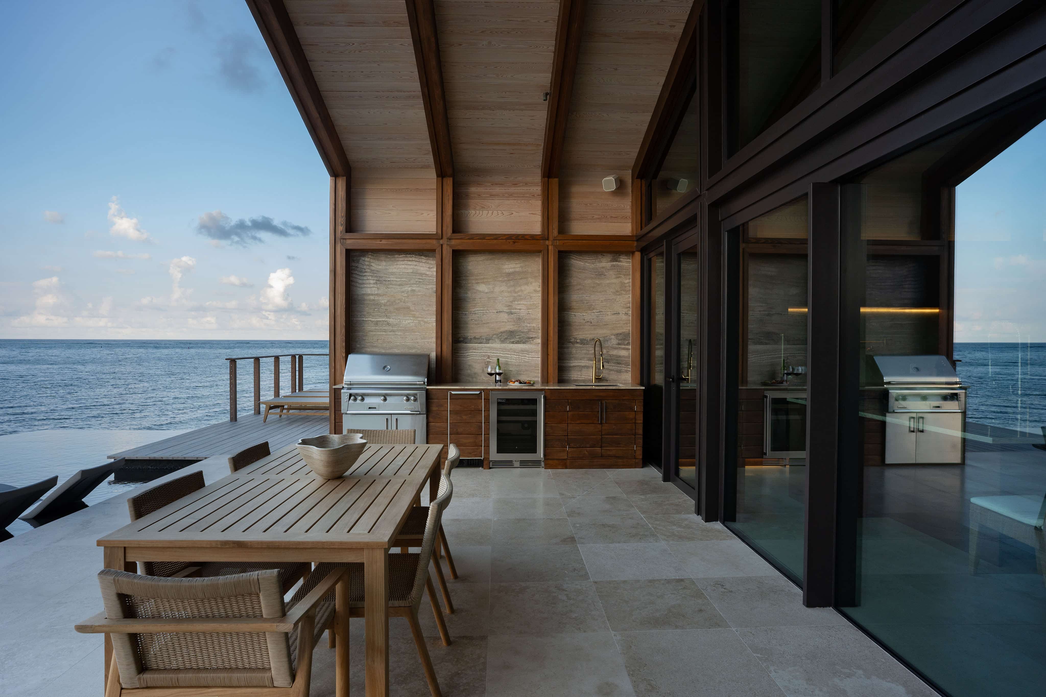 A modern outdoor patio at Ocean Villa, featuring a wooden dining table and chairs overlooking a vast ocean. The space boasts built-in grills and cabinetry beneath a covered structure with large glass doors, all set on a stone-tiled floor for the ultimate luxury experience.