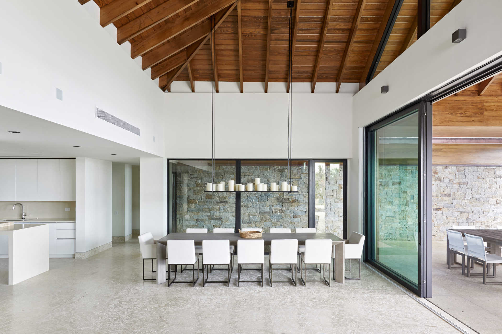 A modern villa dining room with high wooden ceilings features a long dark table surrounded by white chairs. Above the table, a contemporary chandelier with multiple candles hangs. Large sliding glass doors open to an outdoor dining area with a stone wall backdrop.