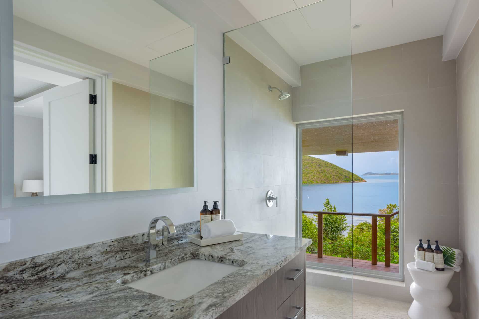 Modern bathroom in Villa DB1 with a large countertop, stainless steel faucet, and mirror. The glass shower door and window offer a serene view of Deep Bay's ocean and greenery, enhancing the tranquil atmosphere.
