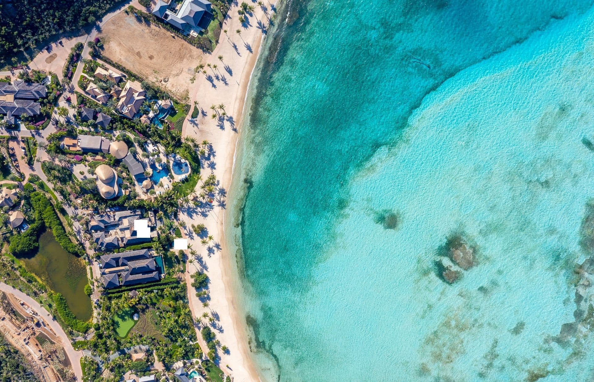 Aerial view of a coastal area with turquoise waters and a sandy beach. An oceanfront resort surrounded by greenery is visible on the left, close to the shoreline. The clear ocean water transitions from light to dark shades of blue and turquoise toward the depths.