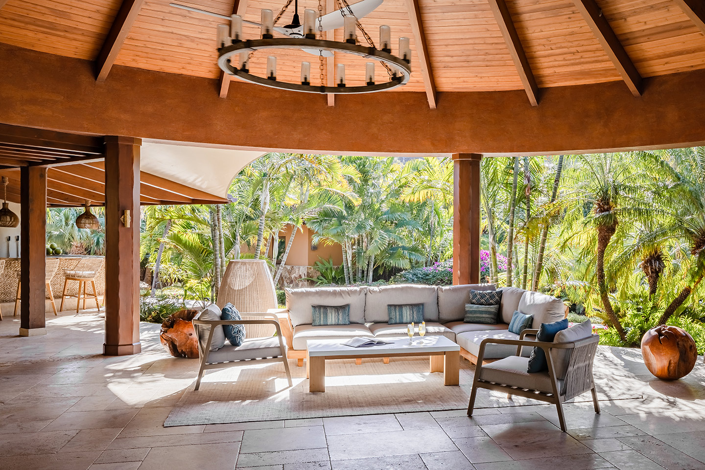A spacious, open-air outdoor lounge area designed like a pavilion at Oil Nut Bay with a circular wooden roof, chandelier, and comfortable seating. Surrounded by lush tropical plants and trees, the cozy couch, coffee table with books, and stylish decor offer a serene and inviting atmosphere.