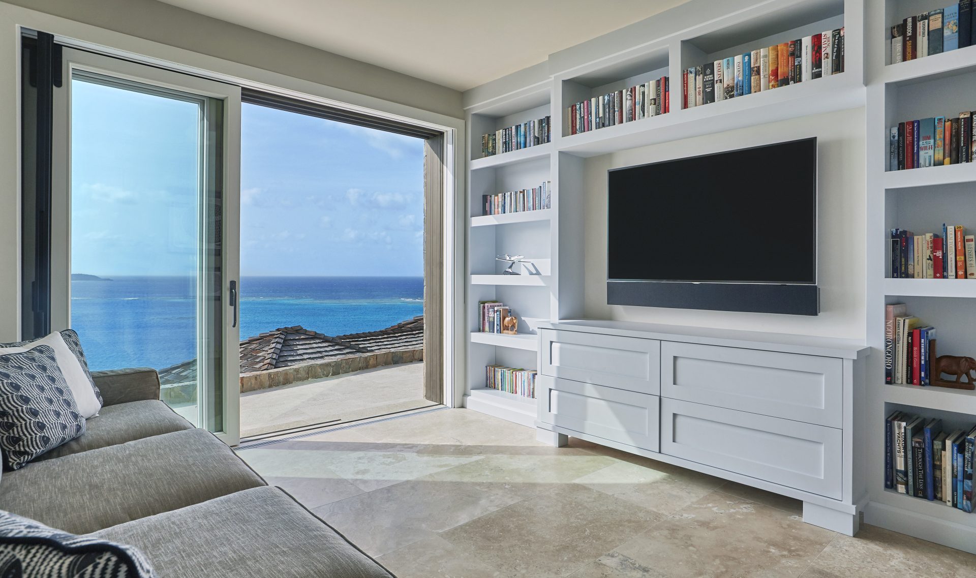A modern living room at the Coral Reef House features built-in bookshelves and a large flat-screen TV on a white entertainment unit. A sliding glass door opens to a balcony with a panoramic view of the ocean and clear blue sky. A grey sofa with patterned cushions faces the TV.