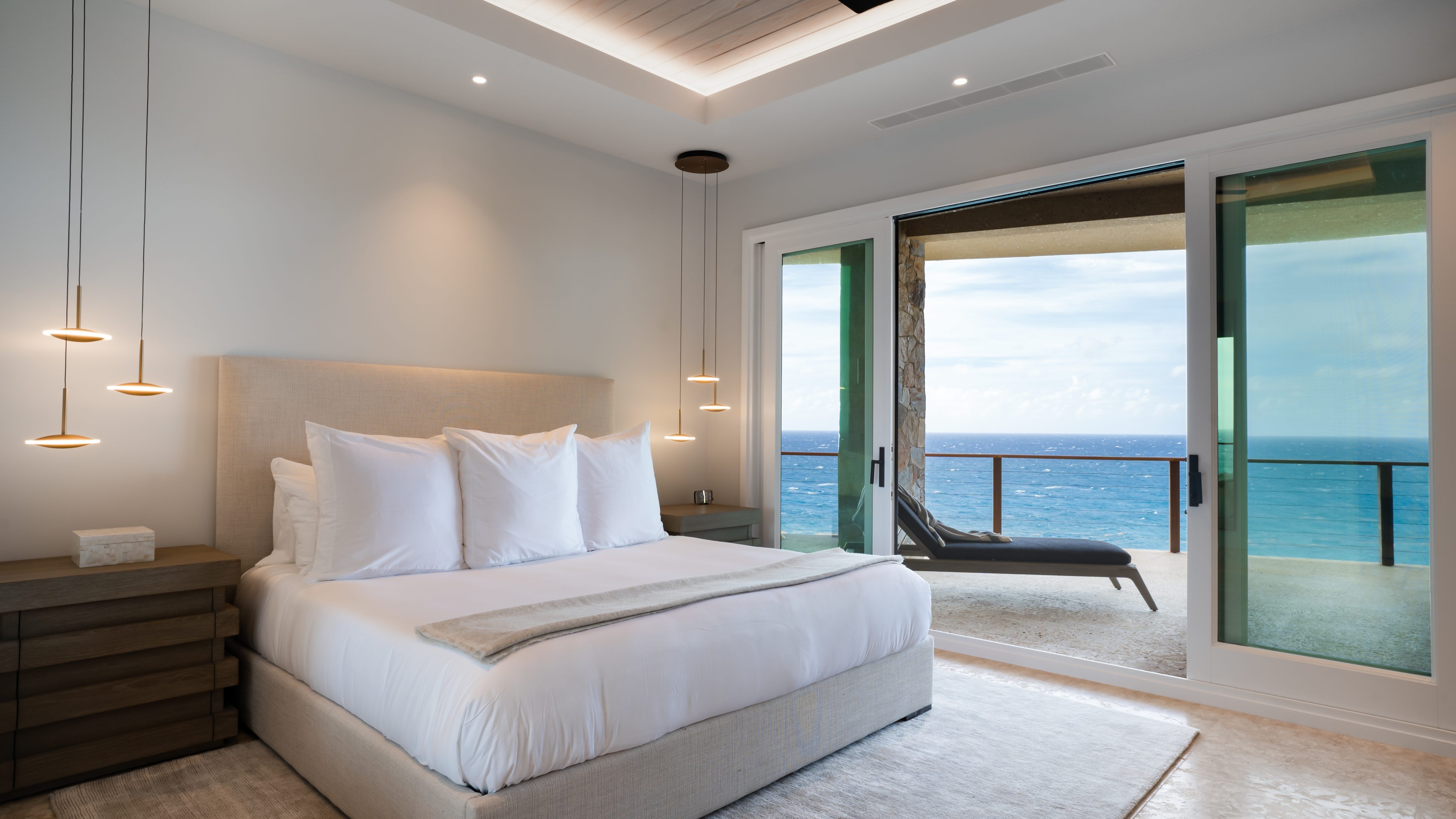Modern bedroom at Hummingbird Villa with a large bed, white bedding, and pendant lights. The room features a sliding glass door opening to a balcony with a lounge chair and a stunning ocean view. Neutral tones and minimalist decor create a serene and inviting atmosphere.
