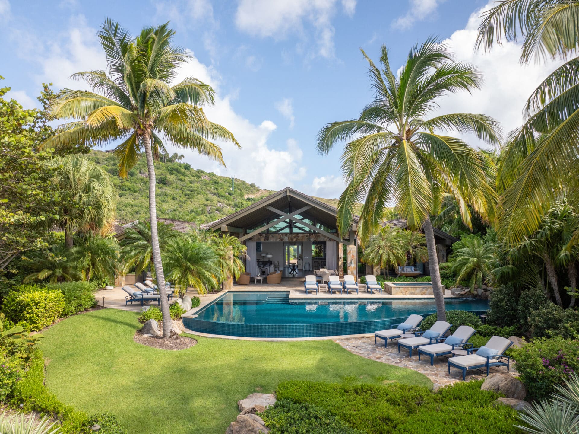 Nonamé Villa is a luxurious tropical escape with an open structure, surrounded by lush greenery and palm trees. It features a spacious outdoor area with a pristine infinity pool, several lounge chairs, and a manicured lawn leading up to the entrance. Blue sky and light clouds overhead.