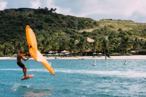 A person wing foiling on blue ocean water near a tropical beach in the British Virgin Islands, holding a bright orange wing. Green hills, palm trees, and umbrellas are visible in the background under a partly cloudy sky.
