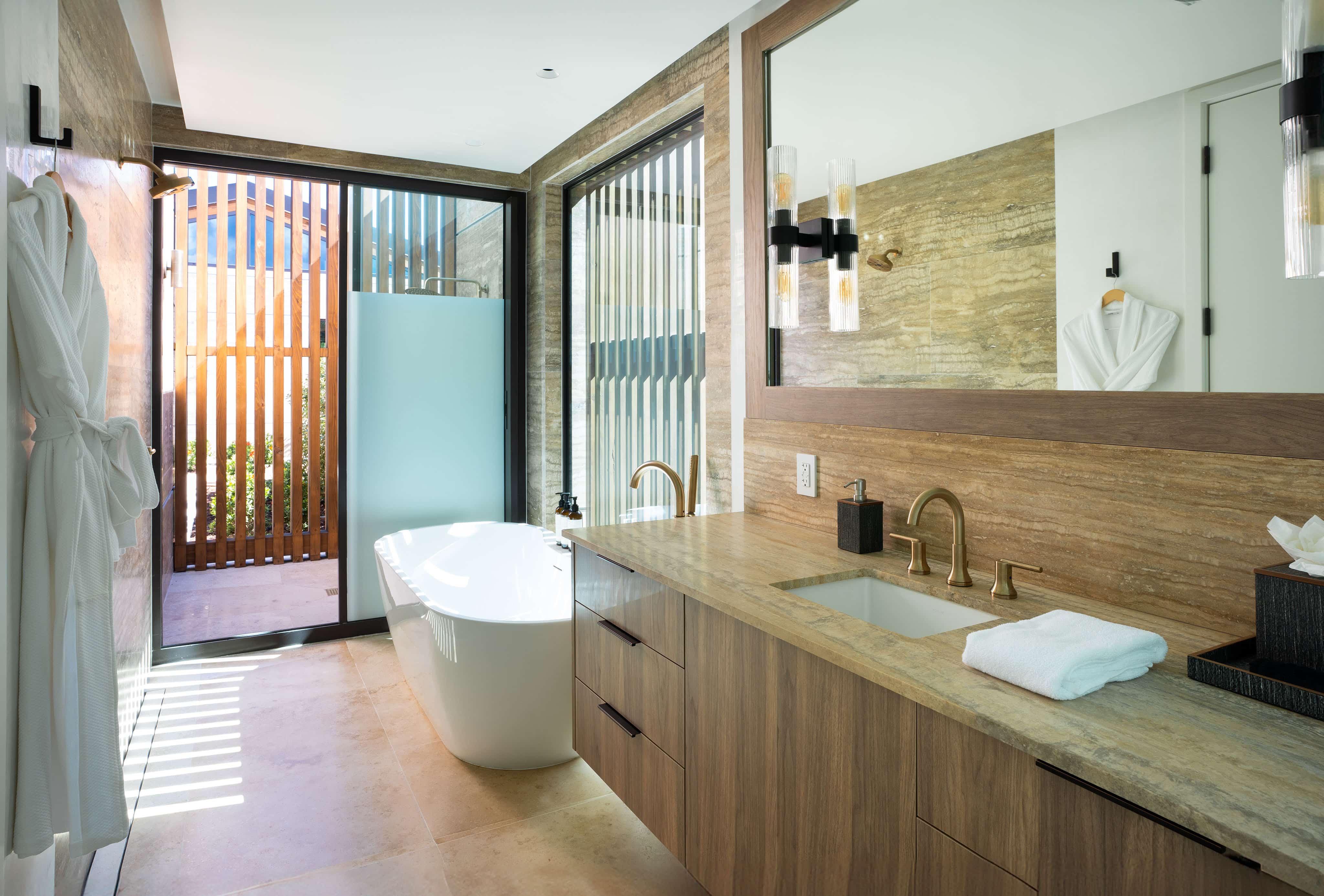 The Ocean Villa's modern bathroom, labeled OV1, showcases natural wood tones with a freestanding bathtub, large mirror, double sink, and glass-enclosed shower. Sunlight streams through a large window with blinds, while towels and a bathrobe hang on the wall.