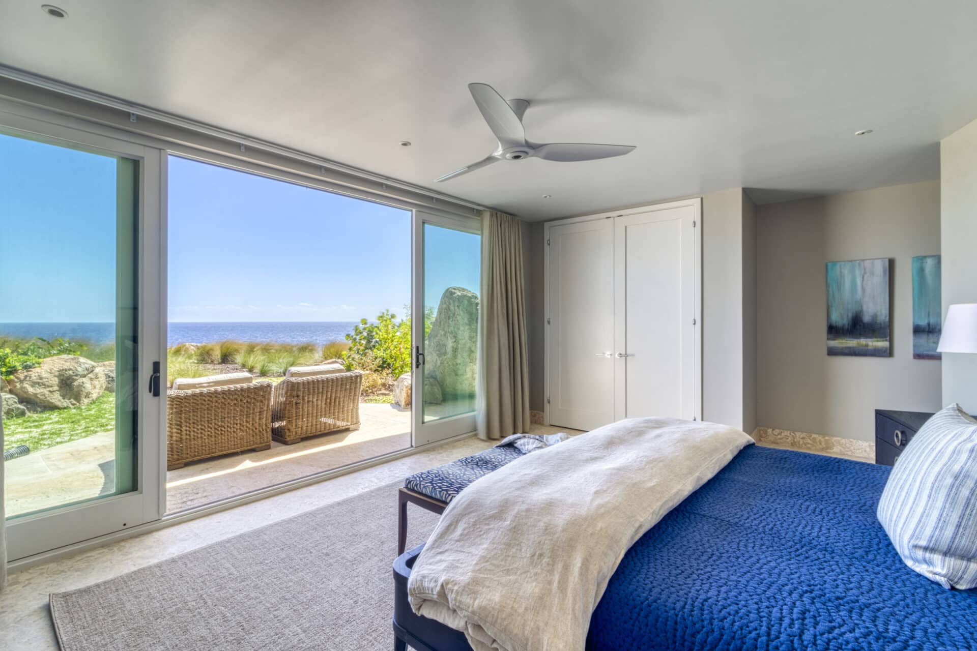 A modern bedroom in Halo Villa with a large sliding glass door leading to an outdoor patio. The room features a bed with a blue blanket, a ceiling fan, and a closet. The patio has two wicker chairs and offers a scenic view of the ocean and greenery.