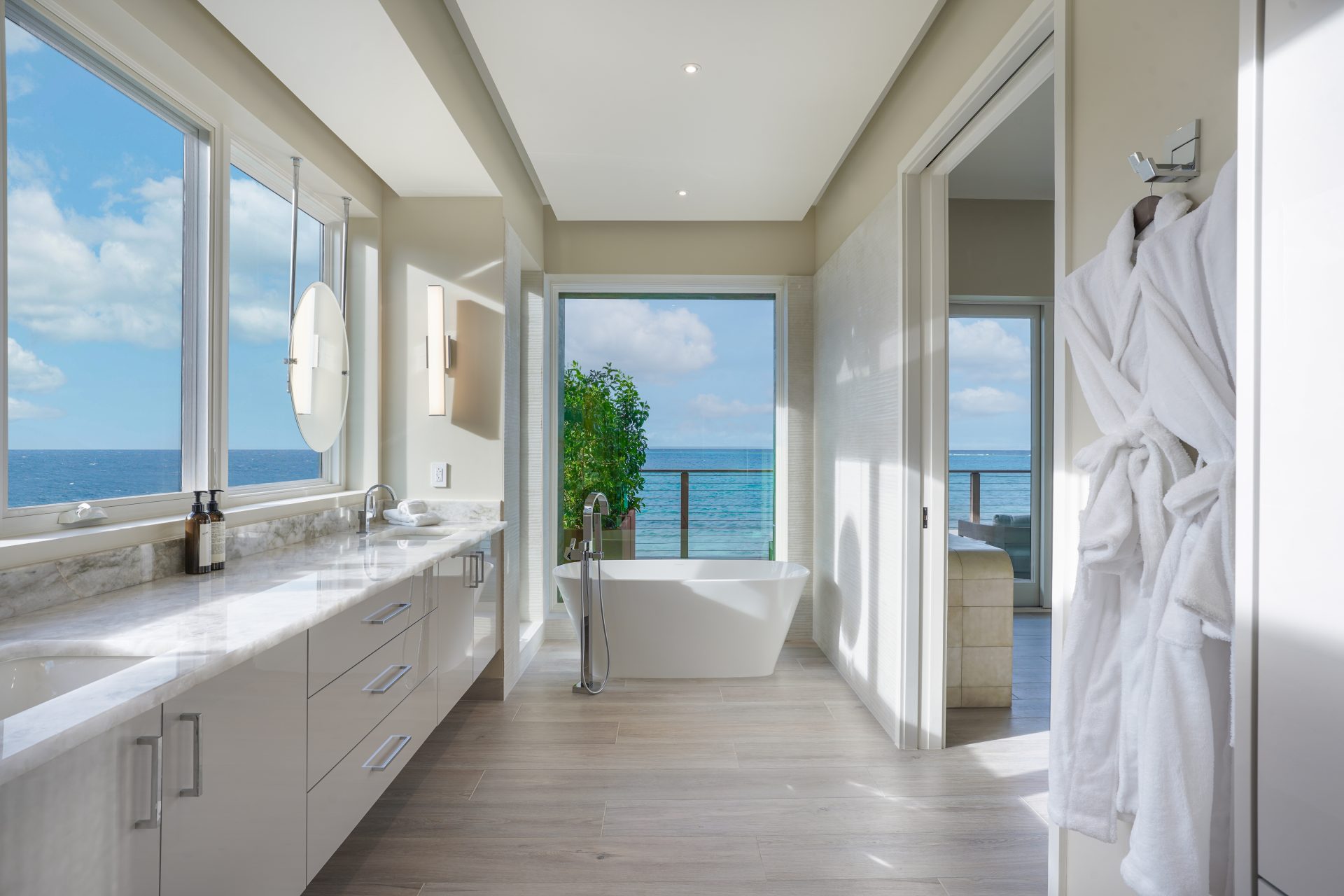 A modern bathroom in Ocean Villa, featuring a large window and balcony offering a stunning ocean view. The room includes a white bathtub, a double sink vanity with marble countertops, and a hanging white bathrobe. The space is bright and airy with light wood flooring, perfect for your OV2 experience.