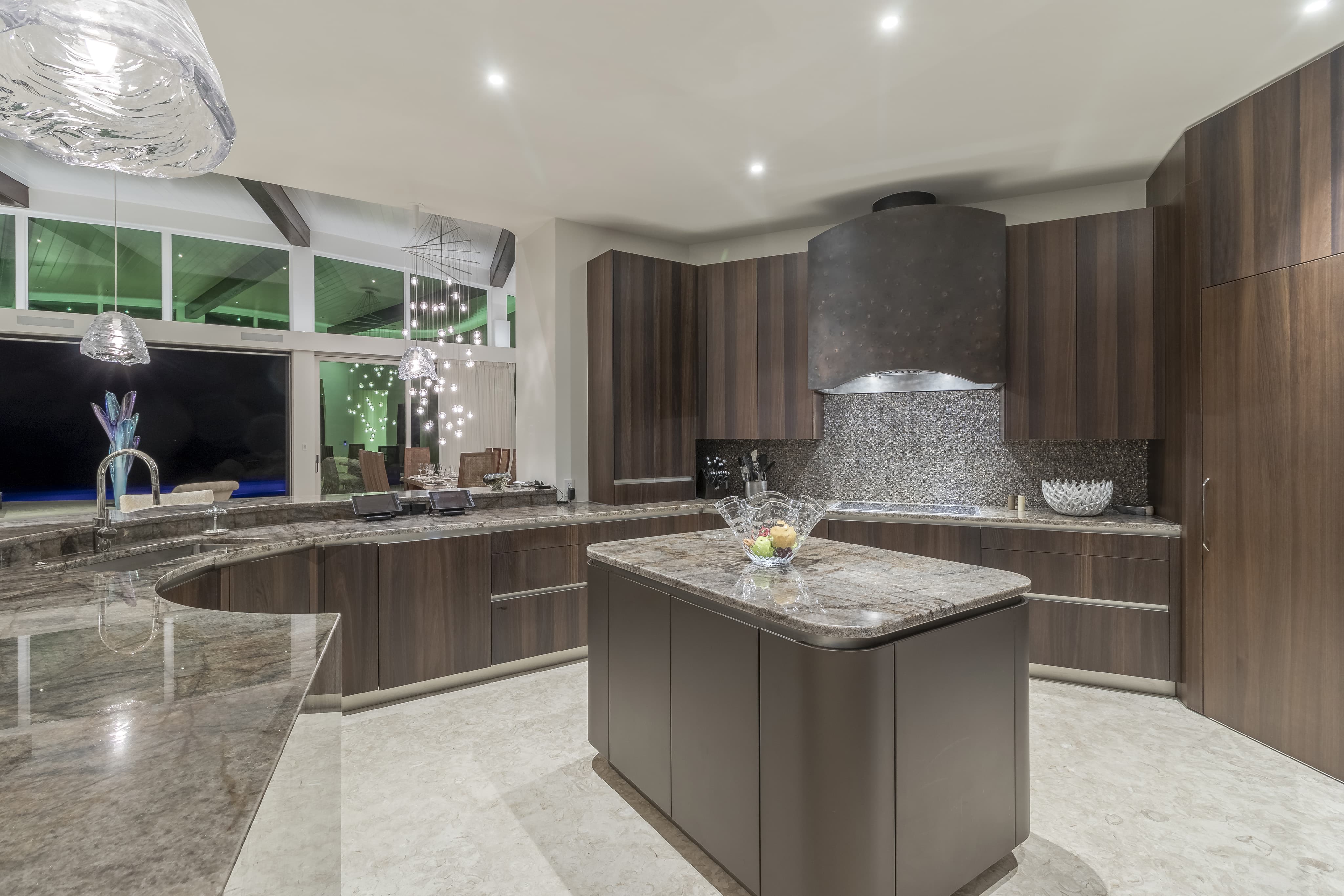 Modern kitchen with sleek dark wood cabinetry and a large marble central island, fitting perfectly within the Halo Villa. The countertops are also marble, and there is a distinctive large black range hood above the stove. A bowl of fruit decorates the island. Ambient lighting creates a cozy atmosphere.