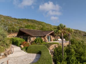 A charming stone villa with a wood roof is nestled in a lush hillside environment. The house features a small patio with chairs, surrounded by dense greenery and palm trees. A pathway lined with hedges and flowers leads to the entrance, under a clear blue sky.