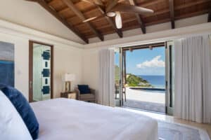 A serene bedroom with a high wooden ceiling and a ceiling fan features a large bed with white linens. Glass doors open to a stunning ocean view with lush green hills and a pool. Light floods the villa, highlighting its airy and relaxing ambiance, giving the room wings to embrace tranquility.