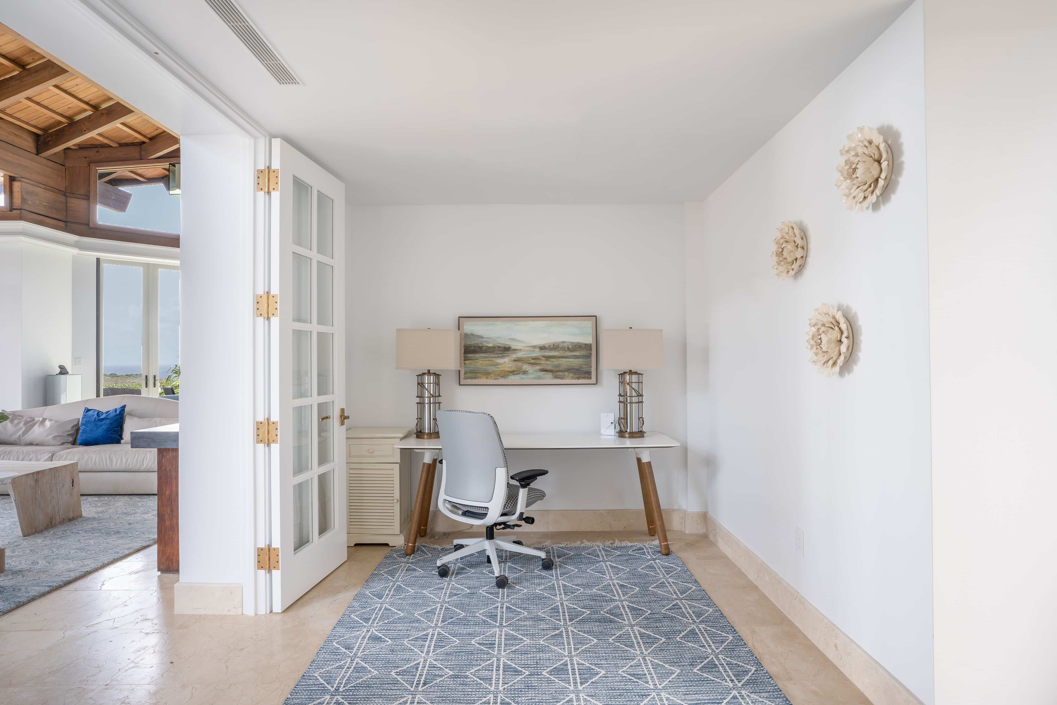 A minimalist home office in a villa, featuring a sleek white desk with a modern office chair, flanked by two beige lamps. A landscape painting hangs above the desk. The room has white walls, a geometric-patterned rug, and three decorative wall pieces. Wings of the adjoining living room are visible.