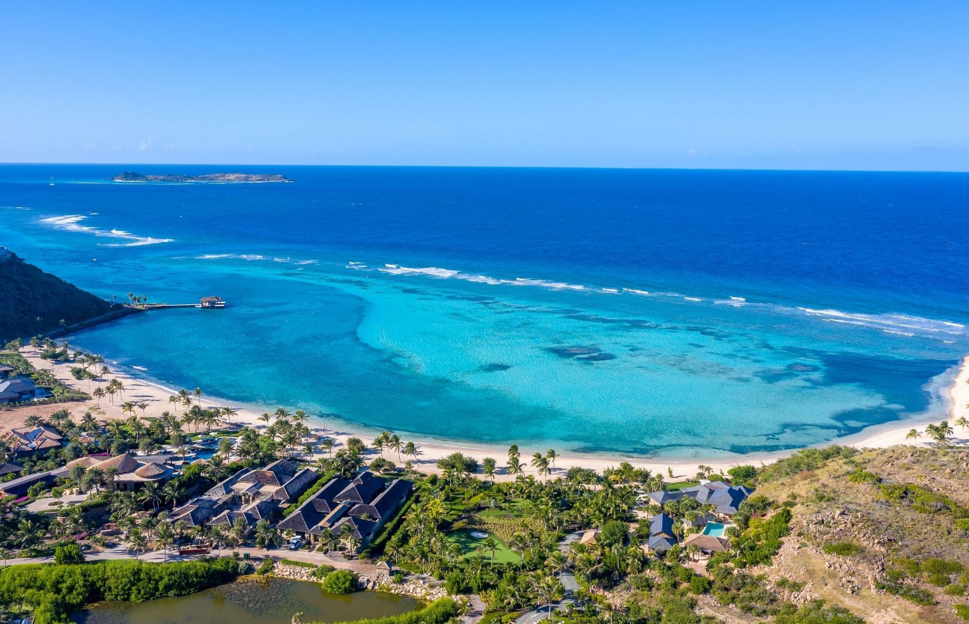 A stunning aerial view of a coastal resort town, one of 2021 travel top destinations, featuring white sandy beaches, clear turquoise water, and lush greenery. The town consists of several buildings and palm trees, with a pier jutting out into the ocean. The horizon shows a small island in the distance.