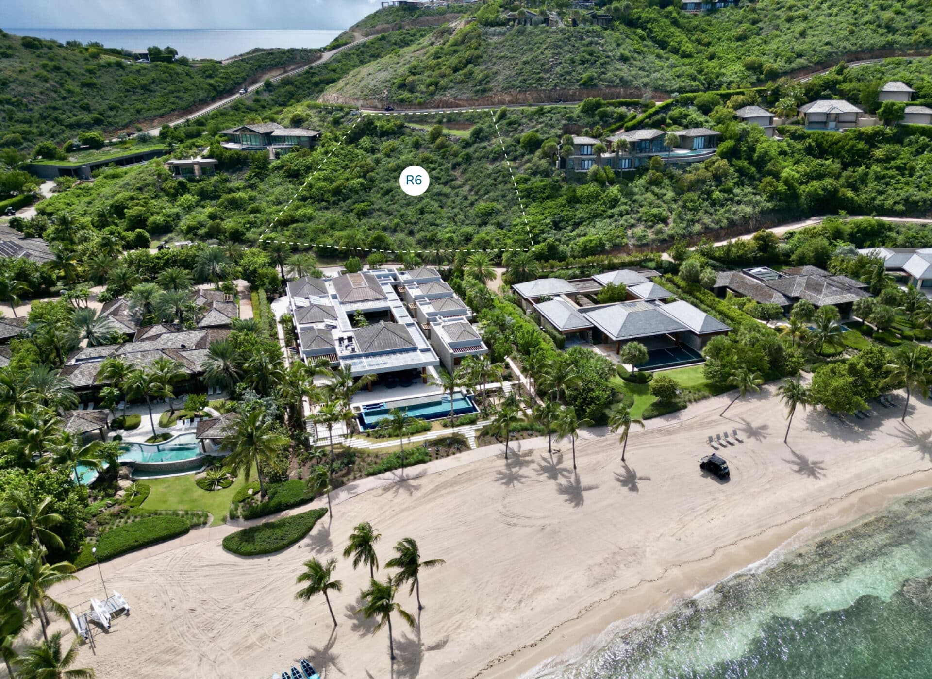 Aerial view of beachfront Ridge Homesite villas surrounded by greenery on a coastal hillside. Large pools, manicured landscapes, and modern architecture define the properties. The sandy beach and calm waters are in the foreground, with lush hills and more homesites in the background.