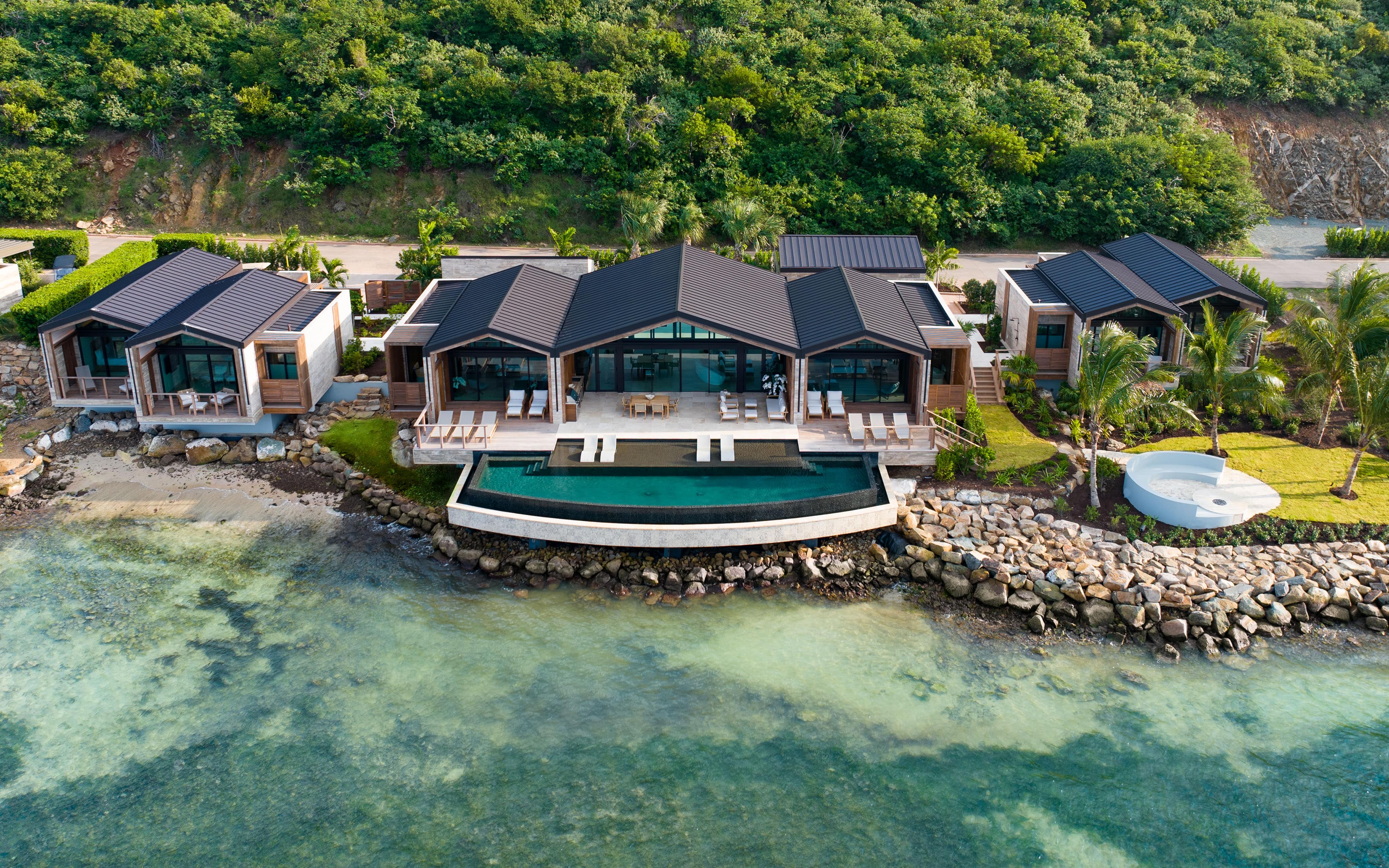 Aerial view of Ocean Villa, a luxury beachfront retreat with modern architecture. The villa features large glass windows, a spacious patio, an infinity pool overlooking the clear ocean water, and lush greenery surrounding the area.