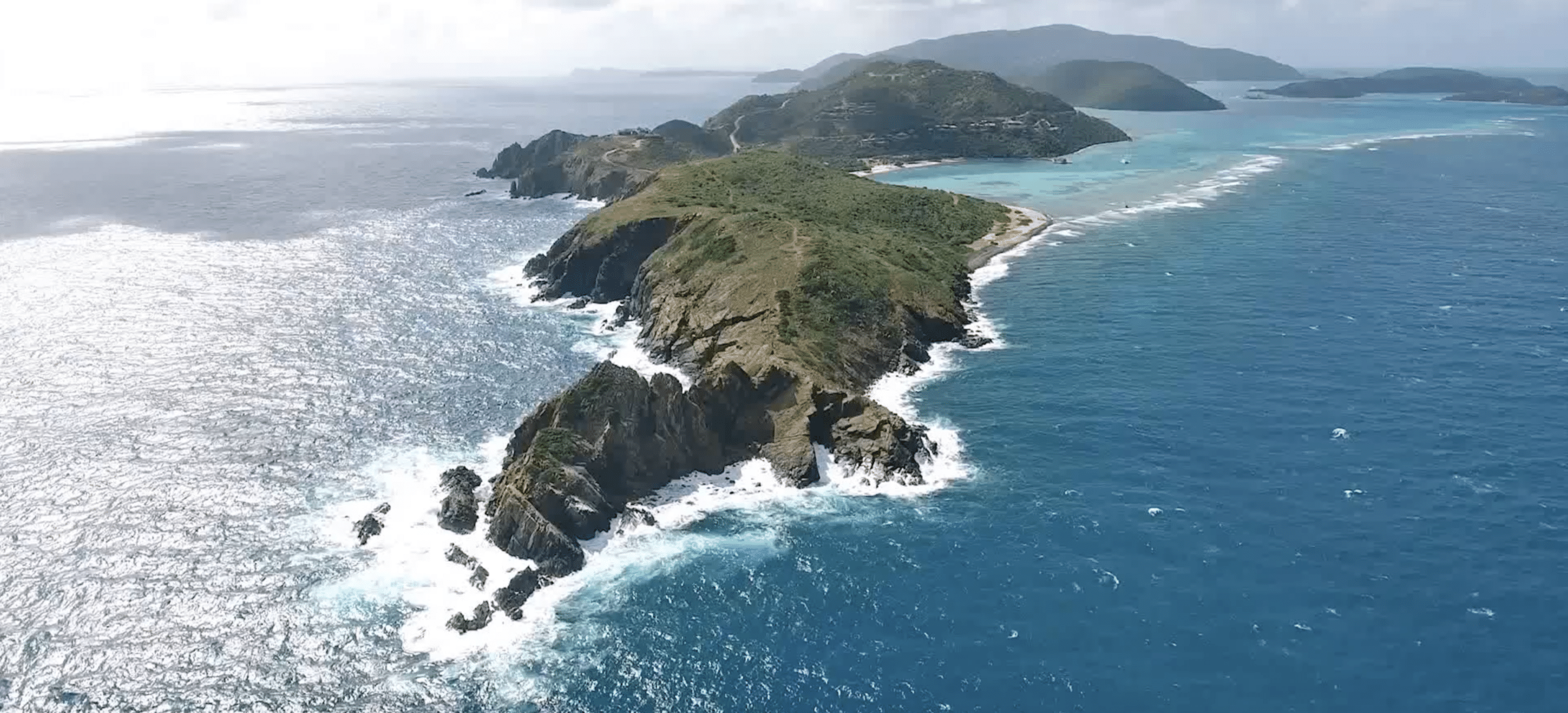 Aerial view of the rugged, narrow Pajaros Point peninsula with steep cliffs and lush green vegetation, surrounded by clear blue ocean waters. Waves crash against the coastline, and distant islands can be seen on the horizon under a partly cloudy sky, marking this scenic neighborhood.
