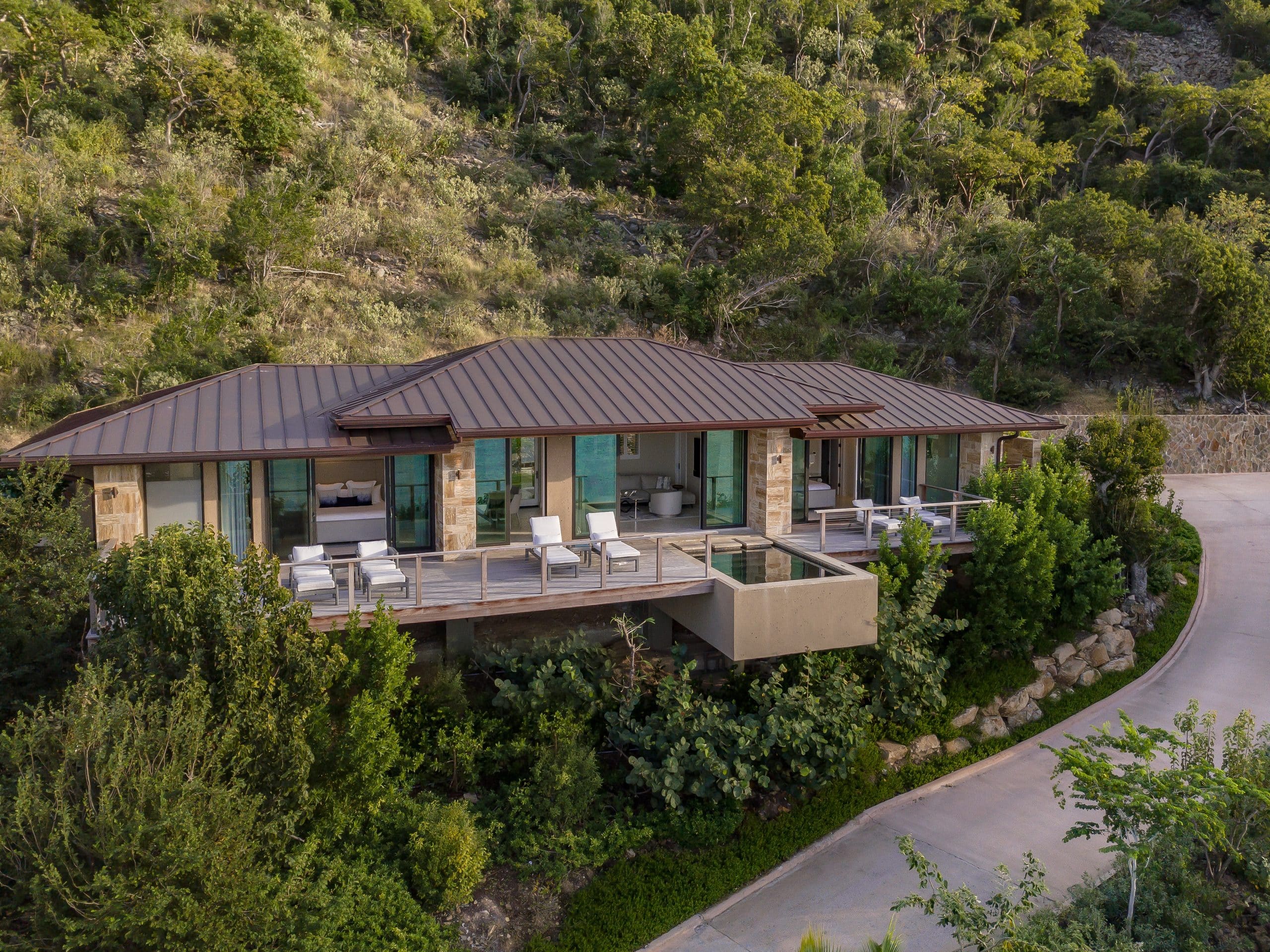A modern villa with a metal roof sits on a hillside surrounded by lush green vegetation. The house features large windows, a spacious deck with outdoor furniture, and a small, elevated pool that extends from the deck. A driveway curves to the right, reminiscent of something MoneyPenny might admire.