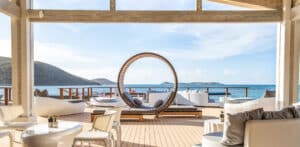 Experience Nova luxury on a beachfront patio with modern white seating, a circular sun bed, and a wooden deck. Overlooking the clear blue ocean and distant islands under a partly cloudy sky, it's ideal for relaxation and enjoying the serene coastal view.