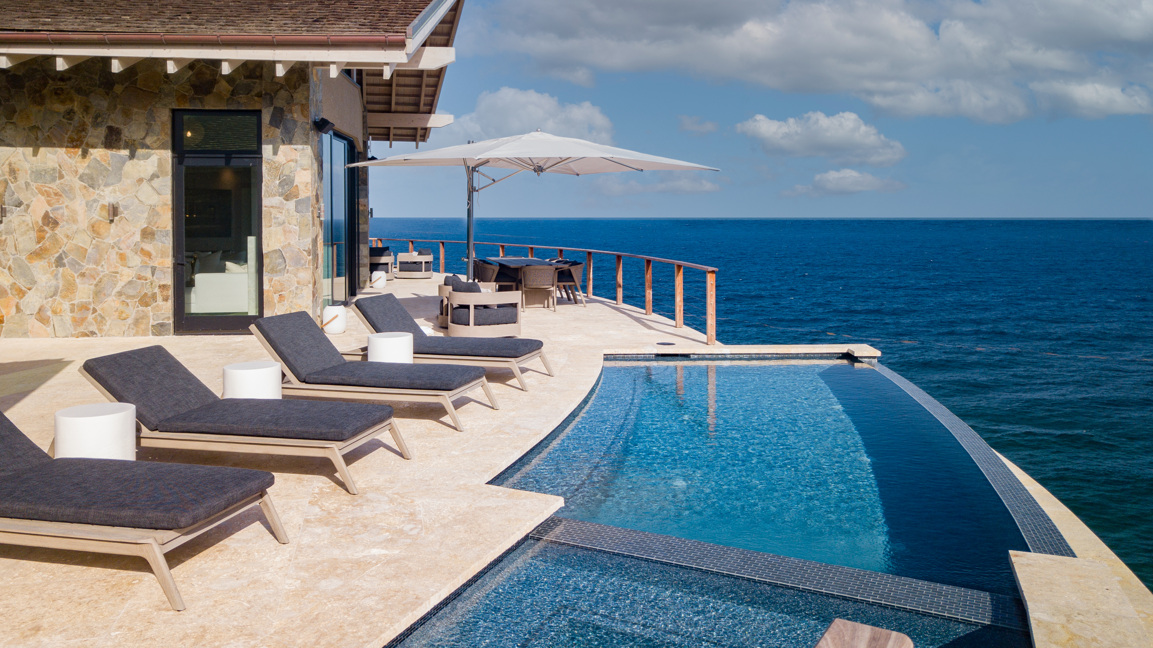 Luxury villa featuring a stone facade, an infinity pool, and four lounge chairs on a sun-drenched terrace overlooking the ocean. An umbrella shades a seating area with a table and chairs, creating a serene and picturesque setting under a partly cloudy sky where hummingbirds occasionally glide by.