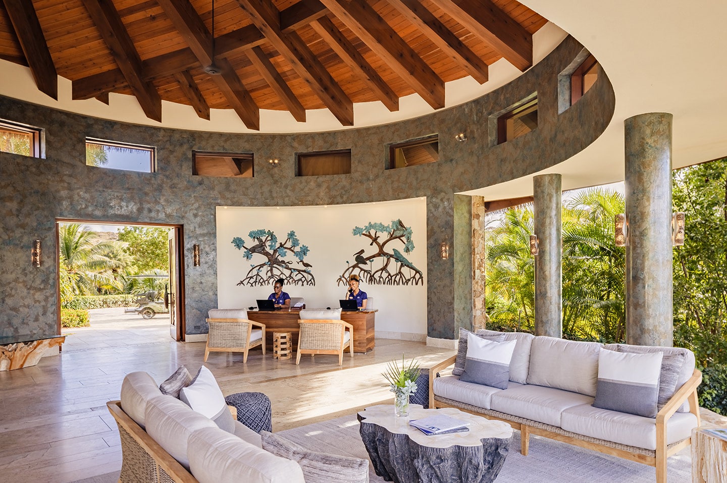 A spacious, open-air lobby at the redesigned Beach Club features a high wooden beamed ceiling and a seating area with cushioned sofas and a unique wooden coffee table. Two receptionists are seated at a wooden desk beneath wall art depicting trees. Large windows and lush greenery are visible.