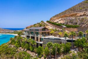 A luxurious hillside villa in Oil Nut Bay overlooks a turquoise ocean, surrounded by lush greenery and palm trees. This multi-story retreat in the British Virgin Islands features large balconies, a private pool, and ample outdoor seating areas under a clear blue sky.