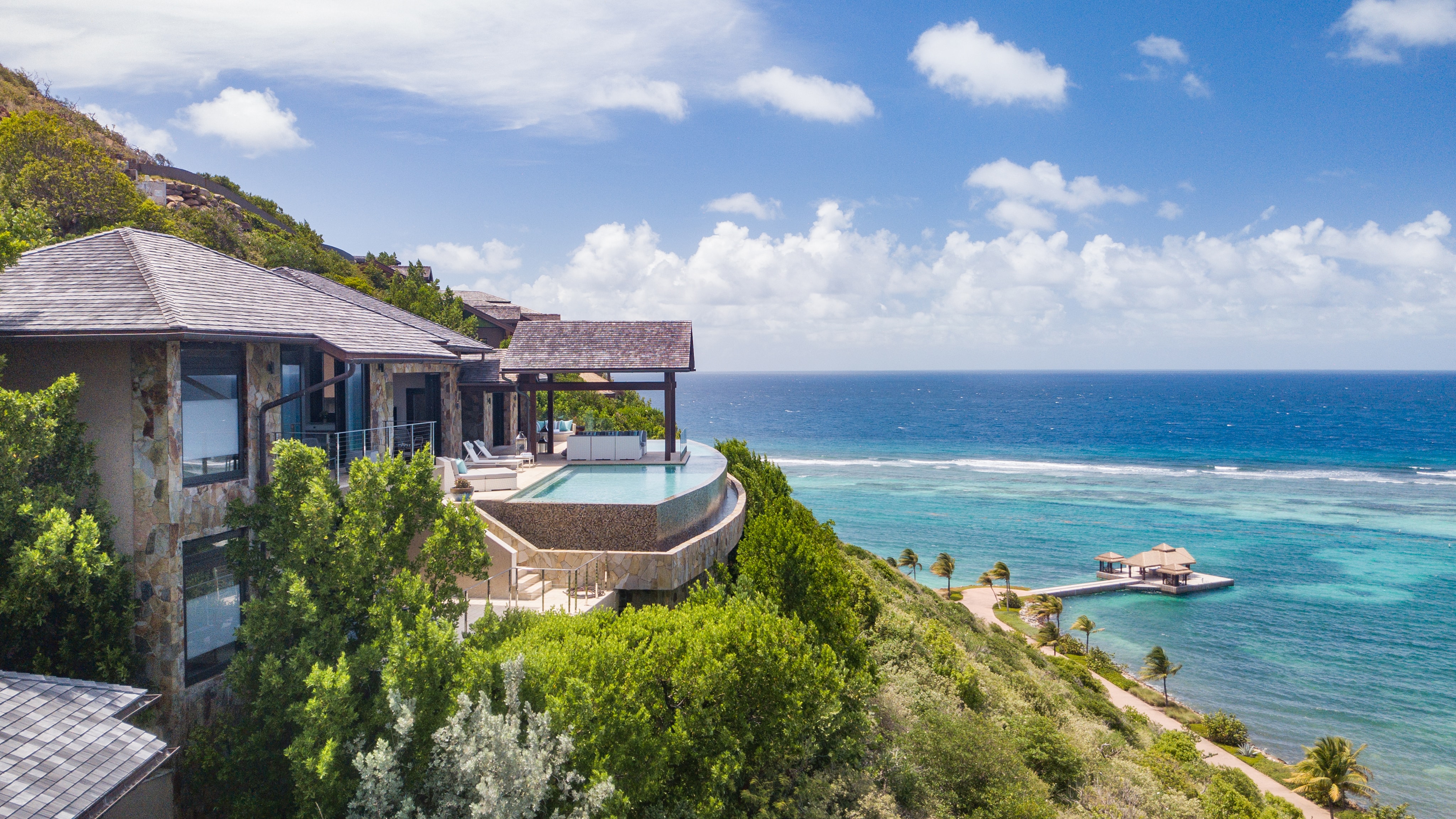 A luxurious villa with a pool overlooks the vast blue ocean of the British Virgin Islands. The villa's architecture blends with the lush green hillside. A cabana rests on a small pier extending into the water, inviting you to escape the cold under fluffy clouds dotting the bright sky.