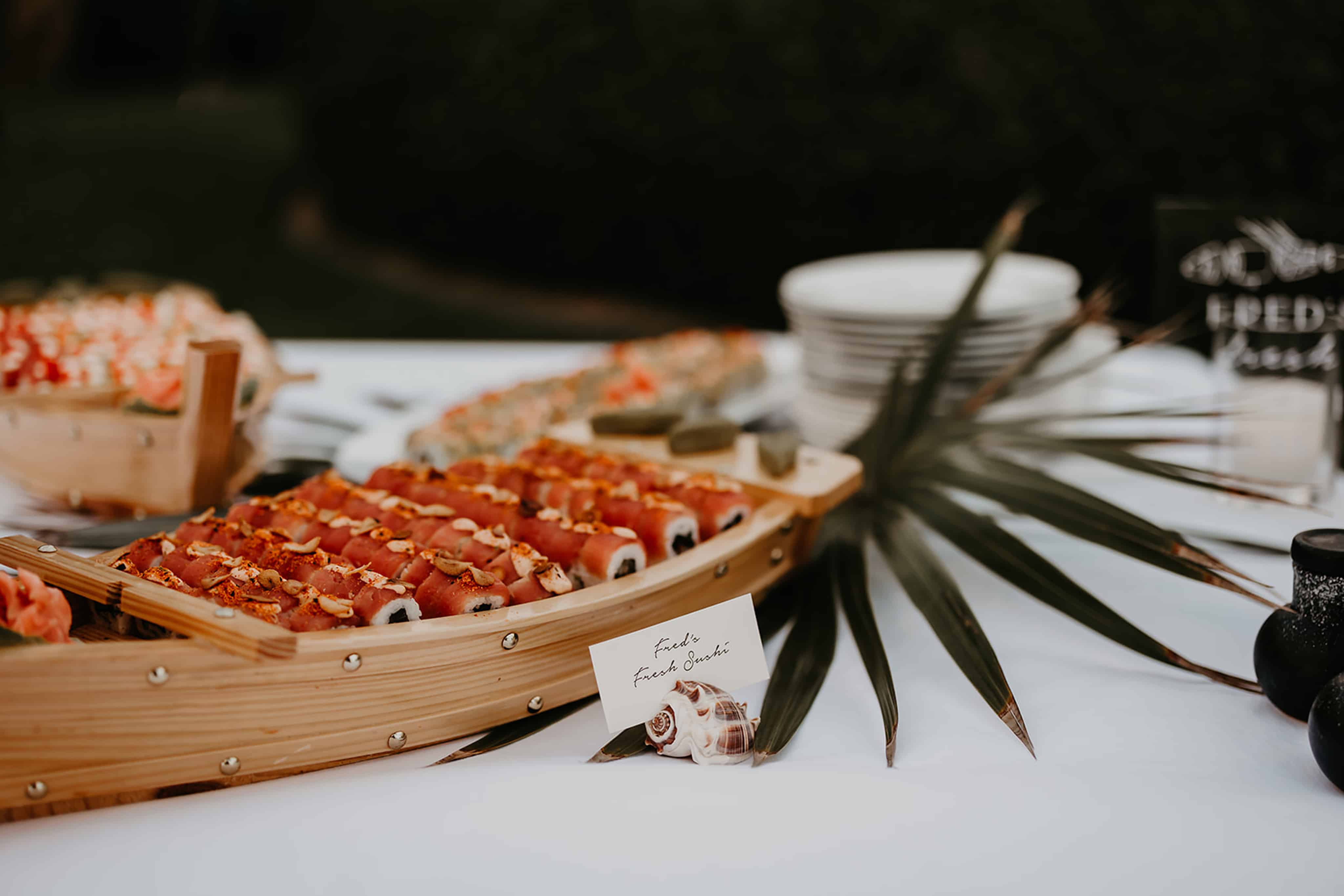 A table displays a variety of sushi artfully arranged on a wooden boat platter, perfect for any BVI Weddings & Events. A palm leaf decorates the setup, and small, neatly stacked plates are visible in the background. A sign in front reads "Fresh Sushi" on a white card with elegant handwriting.