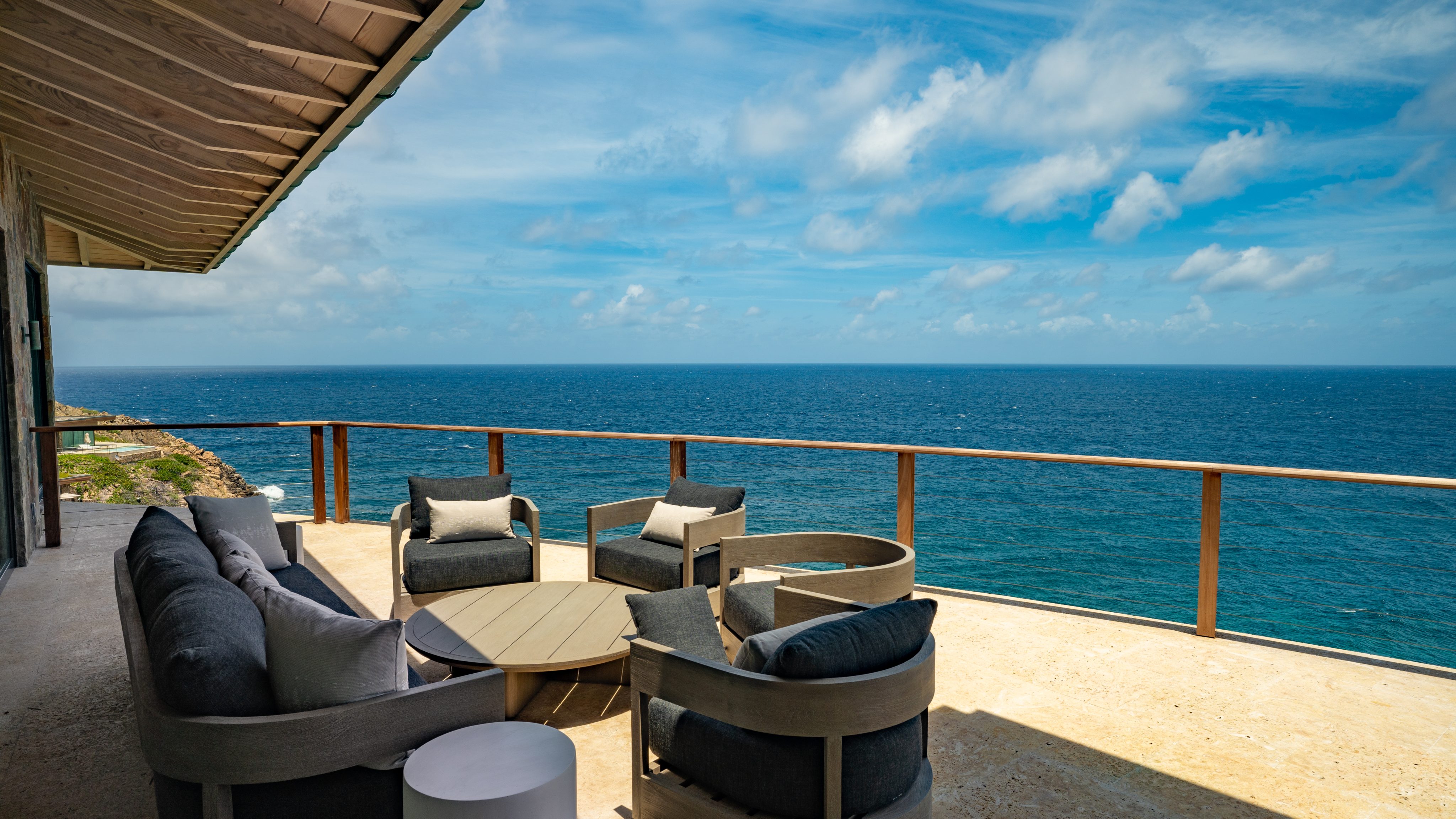 A spacious outdoor terrace at the villa with beige flooring overlooks a vast, clear blue ocean. The area is furnished with grey cushioned chairs, a round wooden coffee table, and a small white side table. A glass railing lines the terrace, providing an unobstructed view of the sea where hummingbirds occasionally flit by.