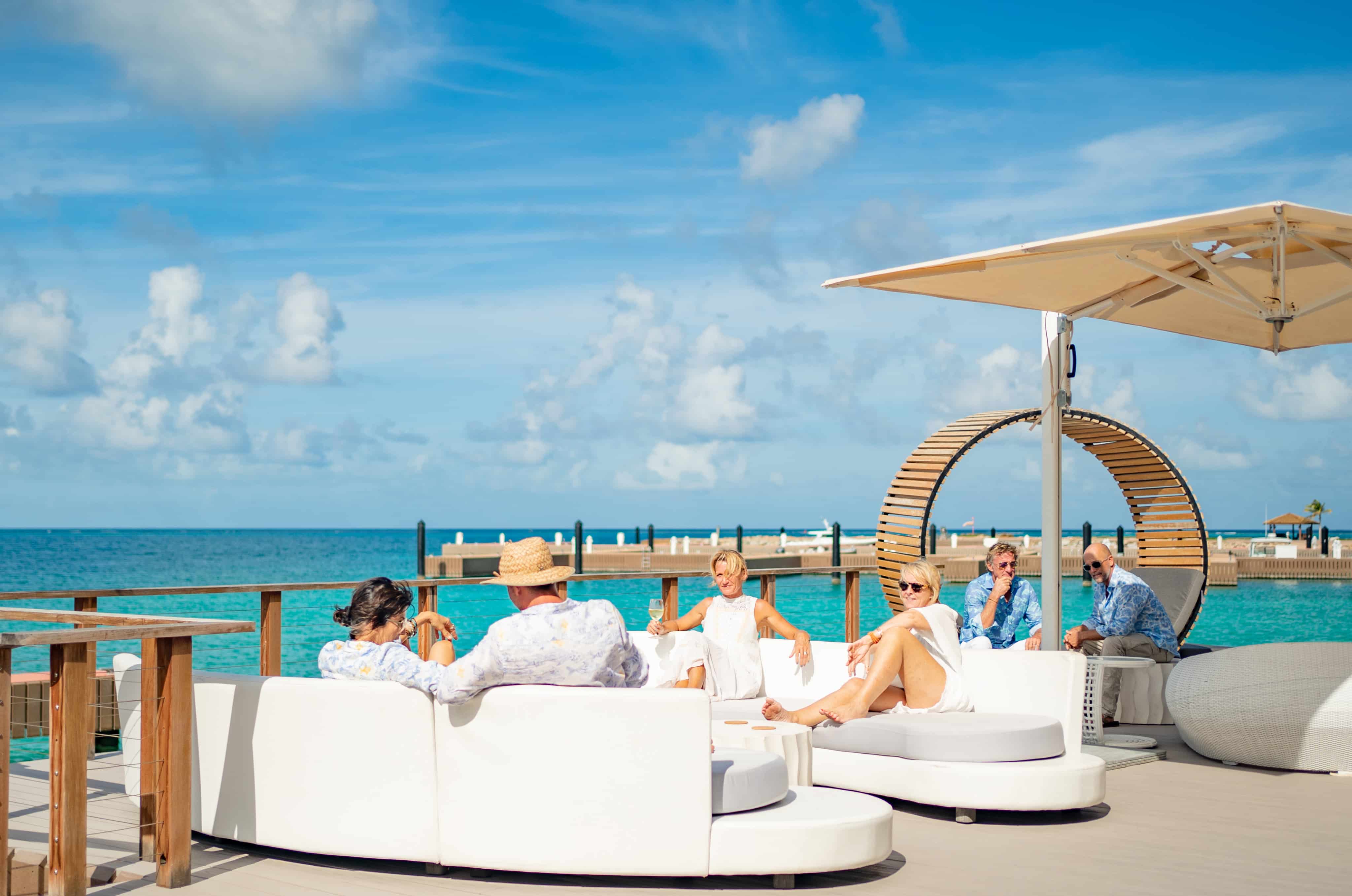 A group of people relaxes on a white Nova outdoor lounge set by the ocean under a sunny blue sky. Some are sitting, while others stand near a modern wooden structure. The scene is vibrant and serene, with a clear view of the water and dock.